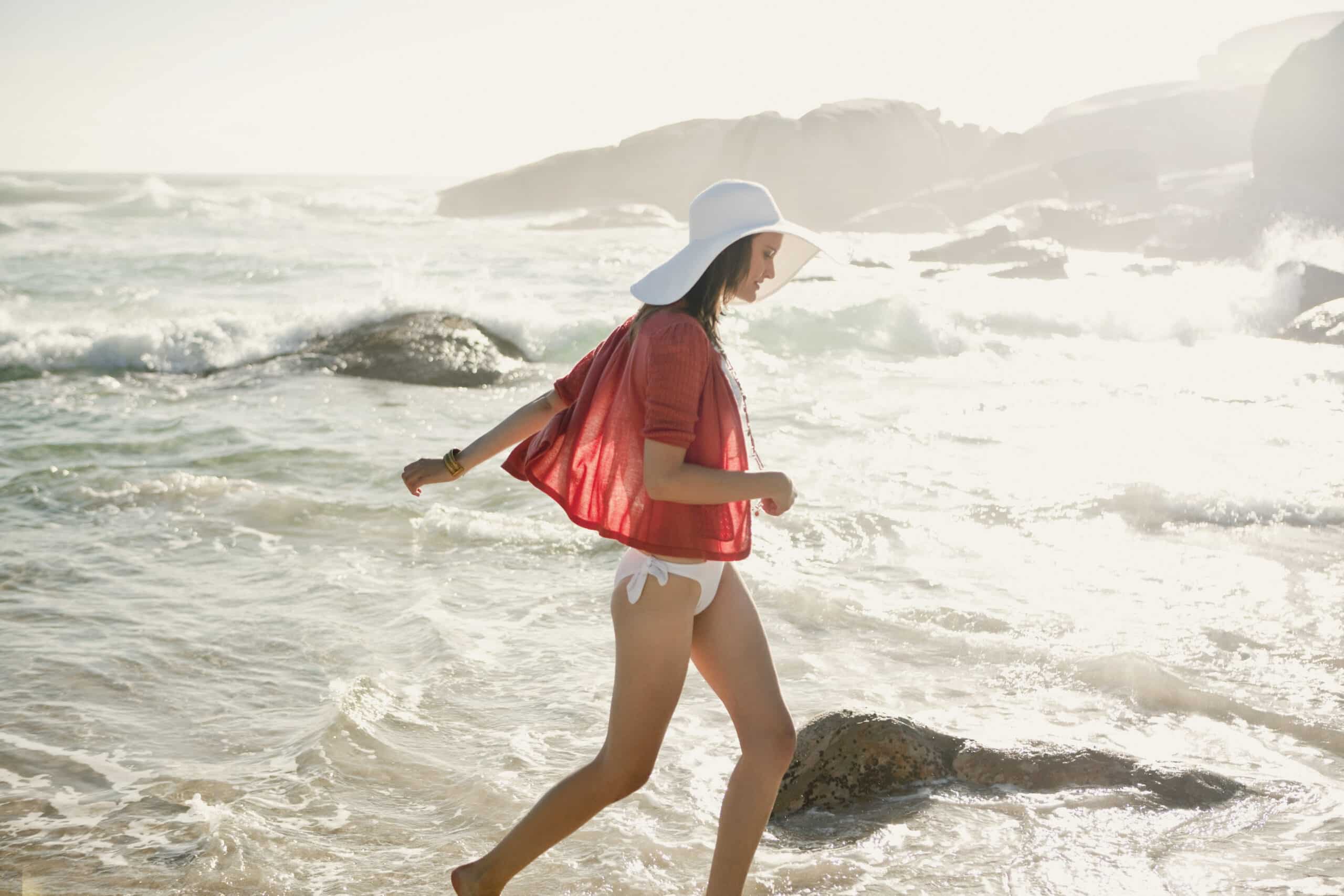 Eine Frau geht an einem sonnigen Strand entlang, in dessen Nähe die Wellen brechen. Sie trägt einen weißen Sonnenhut, einen roten Schal und einen weißen Bikini. Ihr Schatten wirft sich auf den Sand neben ihr. Im Hintergrund sind große Felsen zu sehen. © Fotografie Tomas Rodriguez