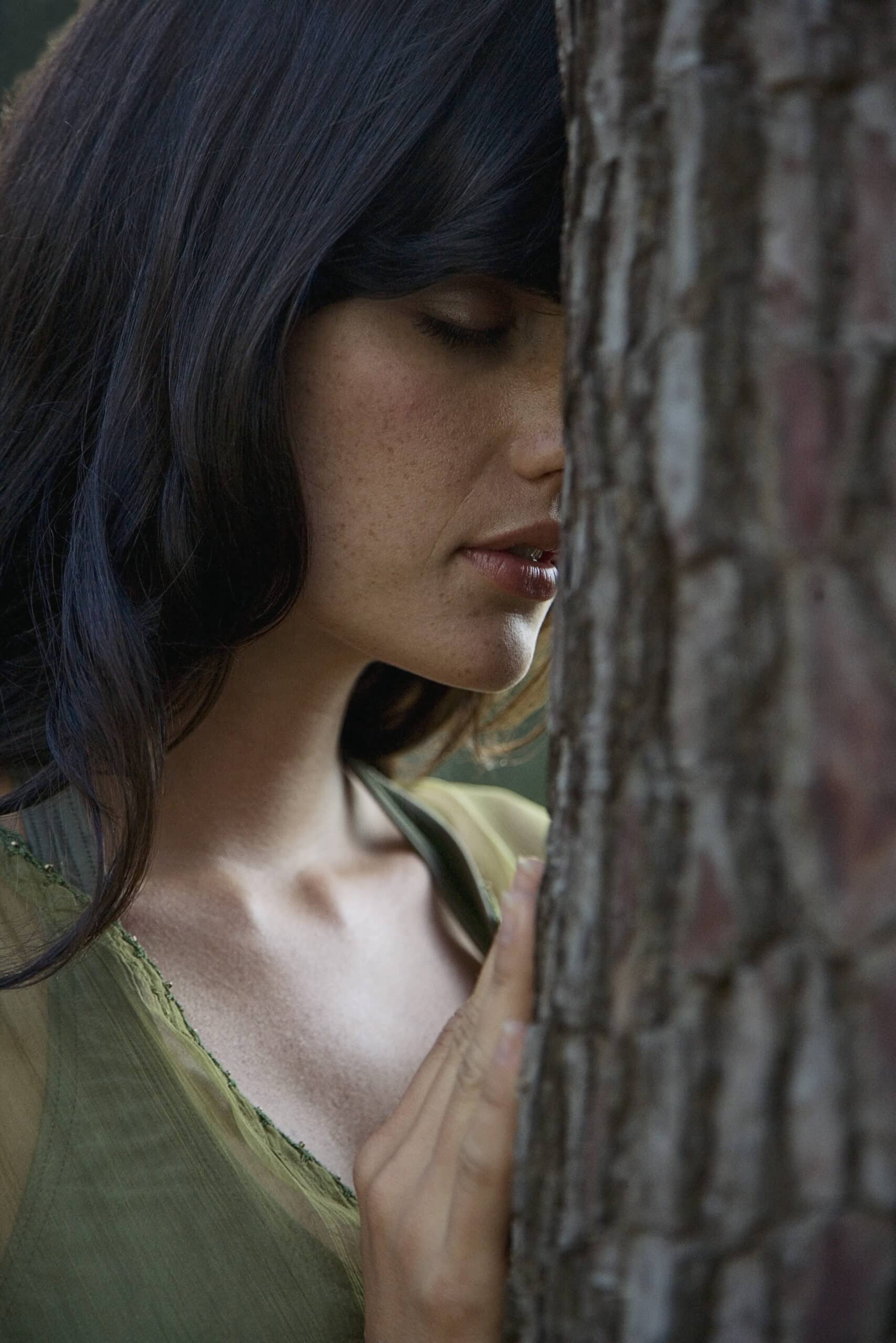 Eine Frau mit dunklem Haar und einem grünen Oberteil steht dicht neben einem Baum, berührt ihn sanft mit der Hand und legt ihre Wange an die Rinde. Ihre Augen sind geschlossen, was einen ruhigen und intimen Moment mit der Natur schafft. © Fotografie Tomas Rodriguez