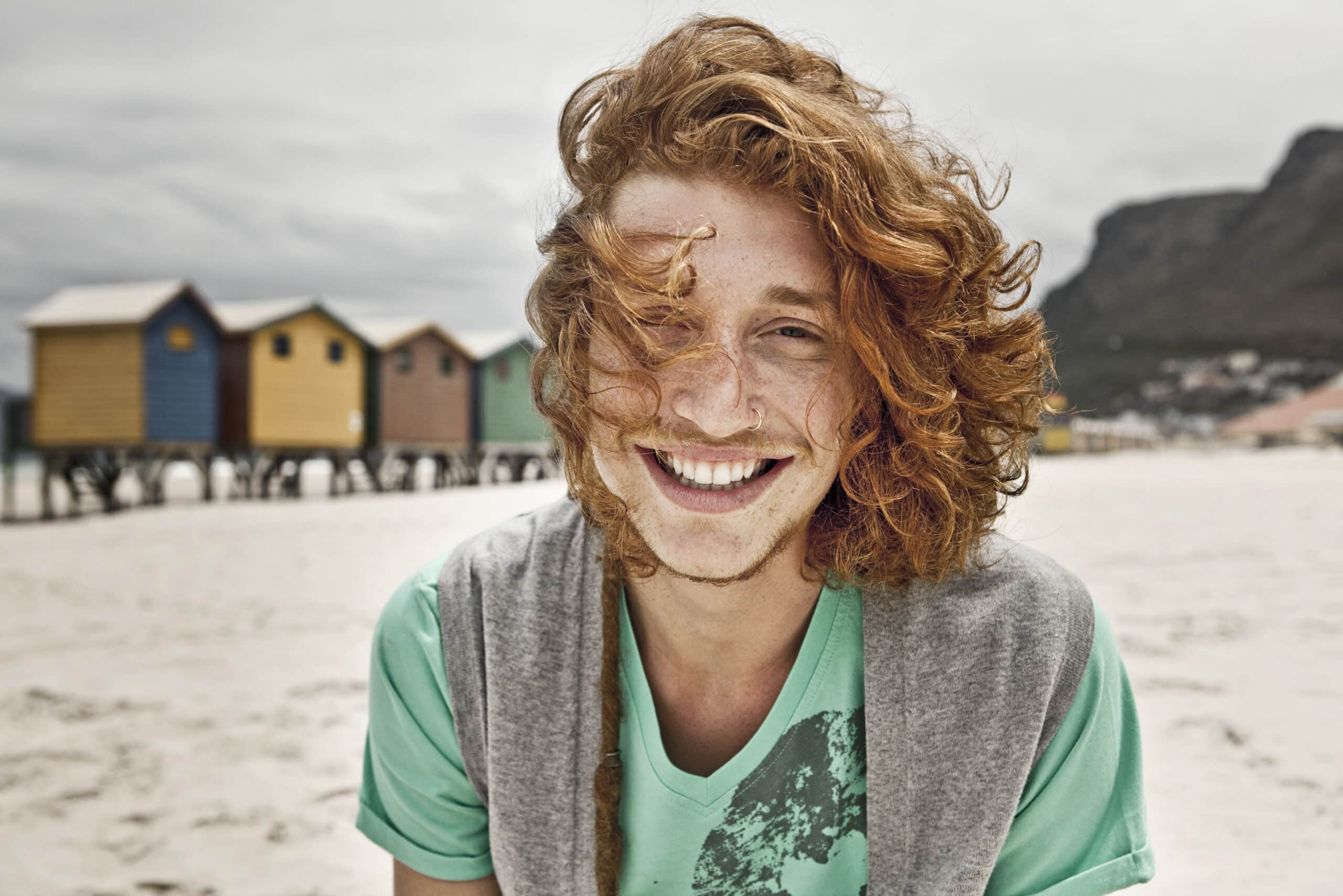 Eine Person mit langen, lockigen roten Haaren und einem breiten Lächeln steht draußen an einem Strand mit bunten Strandhütten und Bergen im Hintergrund. Die Person trägt ein grünes Hemd und einen grauen Pullover über den Schultern. © Fotografie Tomas Rodriguez