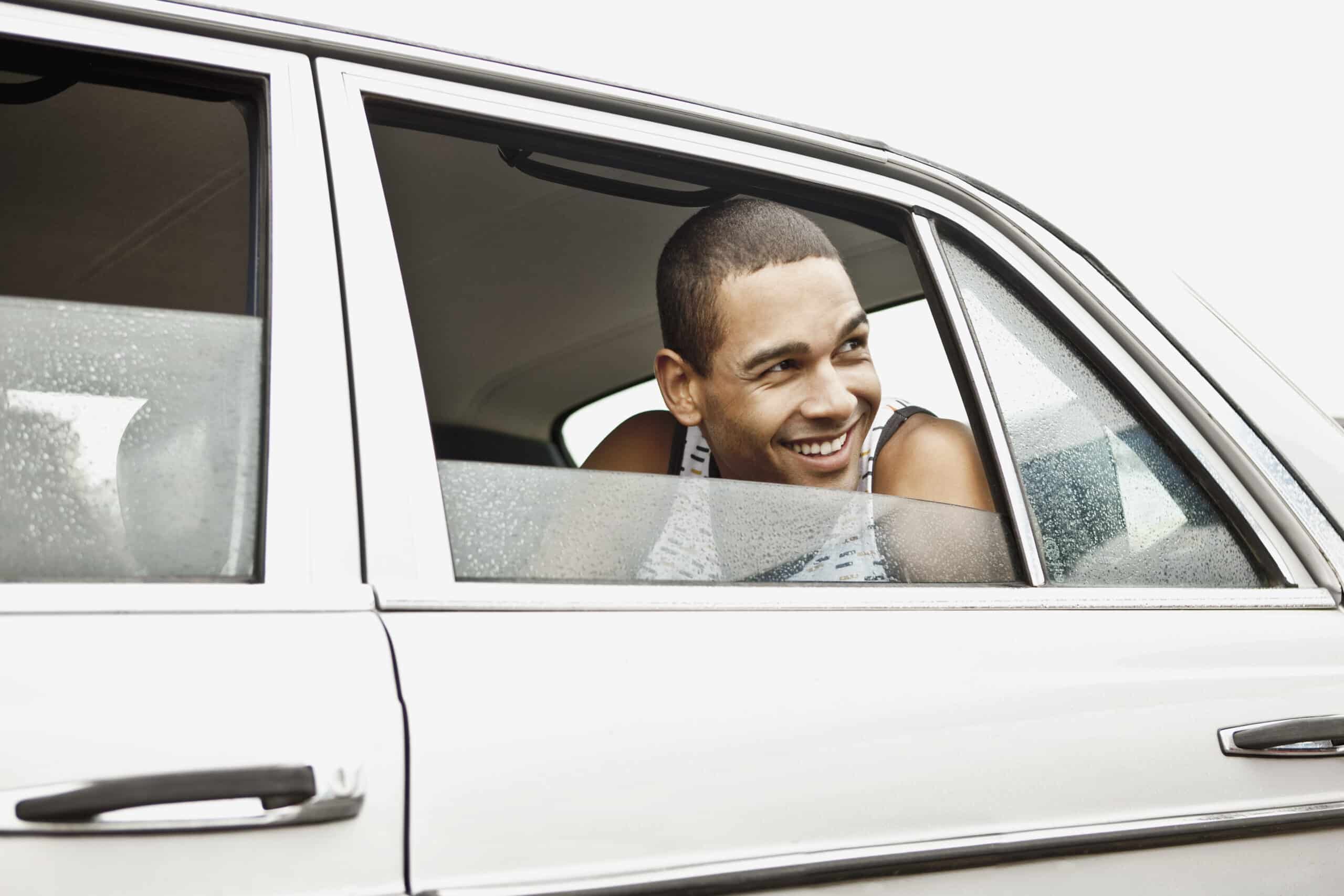 Ein junger Mann mit Buzz Cut lächelt, als er aus dem offenen Fenster eines Autos schaut. Er trägt ein ärmelloses Hemd und scheint den Moment zu genießen. Das Auto ist weiß und hat Wassertropfen an der Außenseite, was darauf hindeutet, dass es vor Kurzem geregnet haben könnte. © Fotografie Tomas Rodriguez