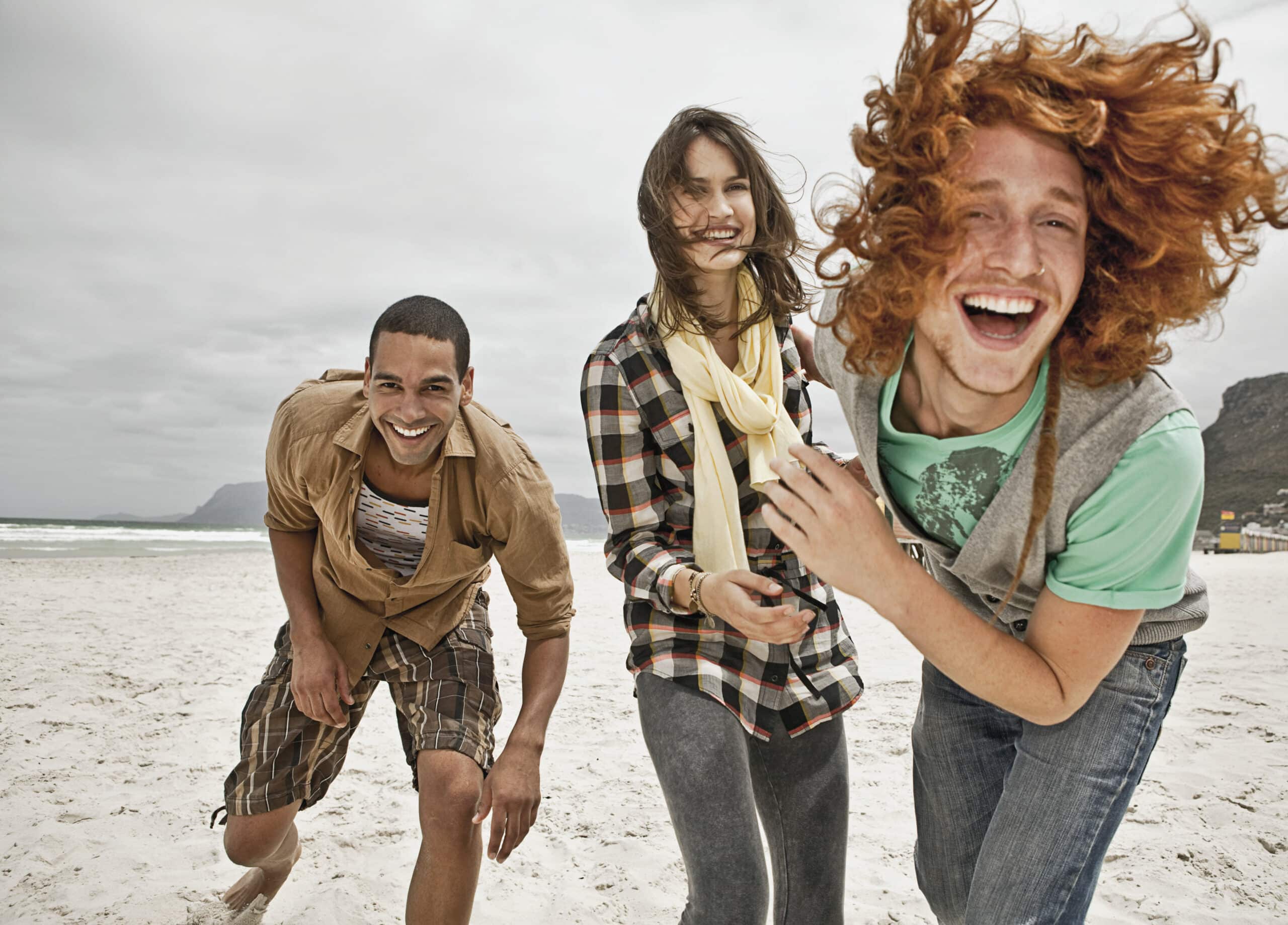 Drei Menschen vergnügen sich am Strand. Sie lachen alle und der Wind weht ihnen durch die Haare. Die Person links beugt sich nach vorne, die Person in der Mitte hält einen Schal und die Person rechts hat lockiges rotes Haar. Im Hintergrund sind das Meer und die Berge zu sehen. © Fotografie Tomas Rodriguez