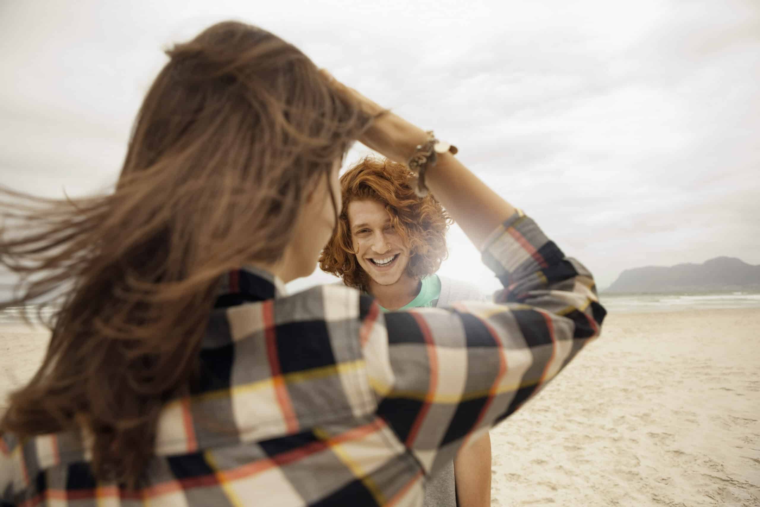 Zwei Personen am Strand; eine mit langen Haaren, die ein kariertes Hemd trägt und von der Kamera abgewandt ist, und eine andere mit lockigem Haar, die in die Kamera lächelt. Im Hintergrund sind Sand und ein bewölkter Himmel mit Bergen in der Ferne zu sehen. © Fotografie Tomas Rodriguez