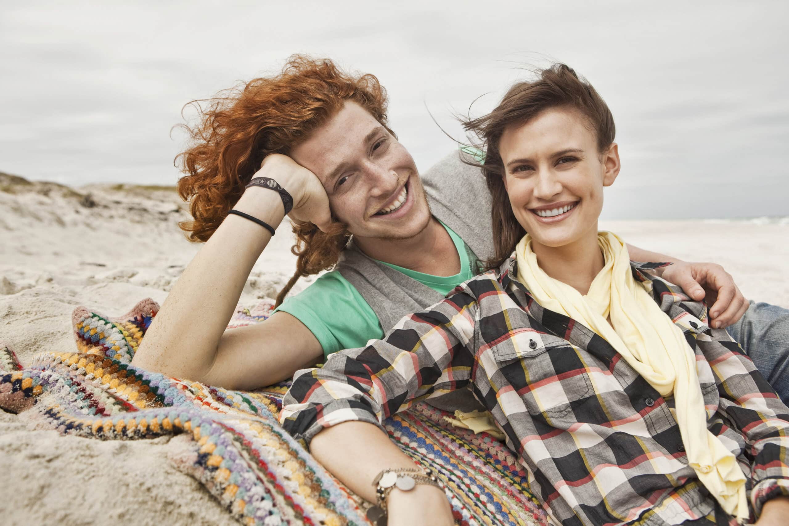 Zwei Menschen liegen auf einer bunten Decke am Strand. Beide lächeln. Die Person links hat rote Locken und ein grünes Hemd und stützt den Kopf auf eine Hand. Die Person rechts hat braune Haare, trägt ein kariertes Hemd und einen gelben Schal. © Fotografie Tomas Rodriguez