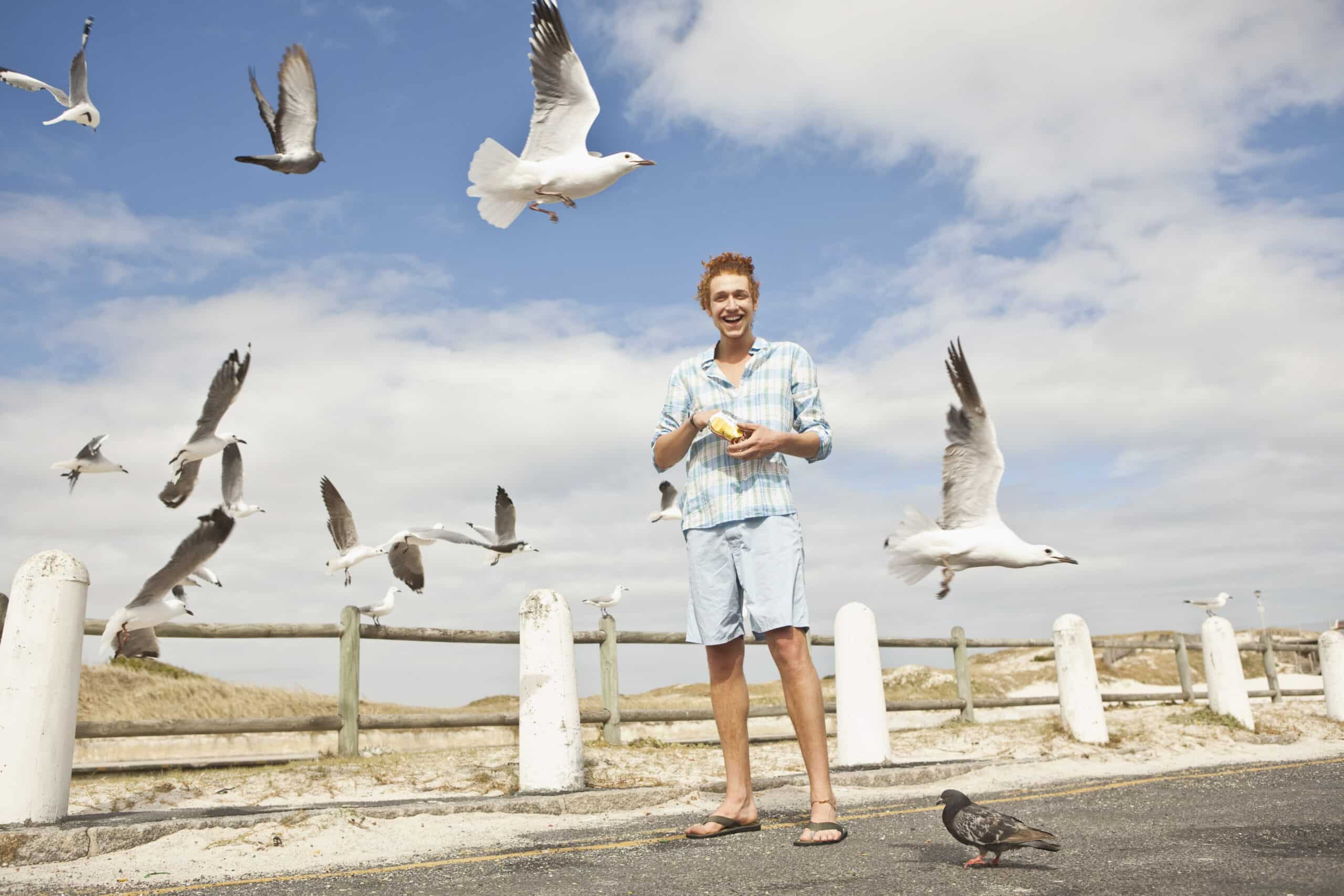 Eine Person mit kurzen Haaren steht auf einem Strandweg und füttert Möwen mit Brotkrümeln. Der Himmel ist teilweise bewölkt und mehrere Möwen fliegen herum. Die Person trägt ein hellblaues Hemd, hellblaue Shorts und Flip-Flops und sieht fröhlich aus. © Fotografie Tomas Rodriguez
