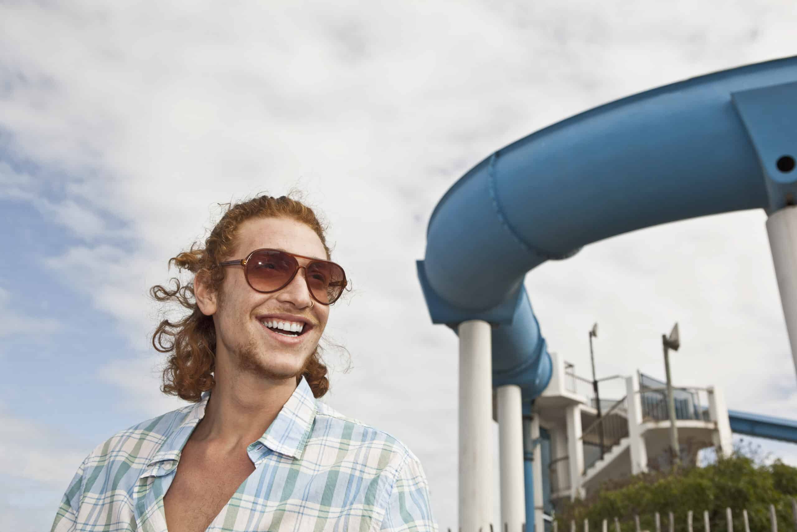 Eine Person mit langen lockigen Haaren, Sonnenbrille und kariertem Hemd lächelt unter einem teilweise bewölkten Himmel. Im Hintergrund ist eine große blaue Wasserrutsche mit weißen Stützen zu sehen. © Fotografie Tomas Rodriguez