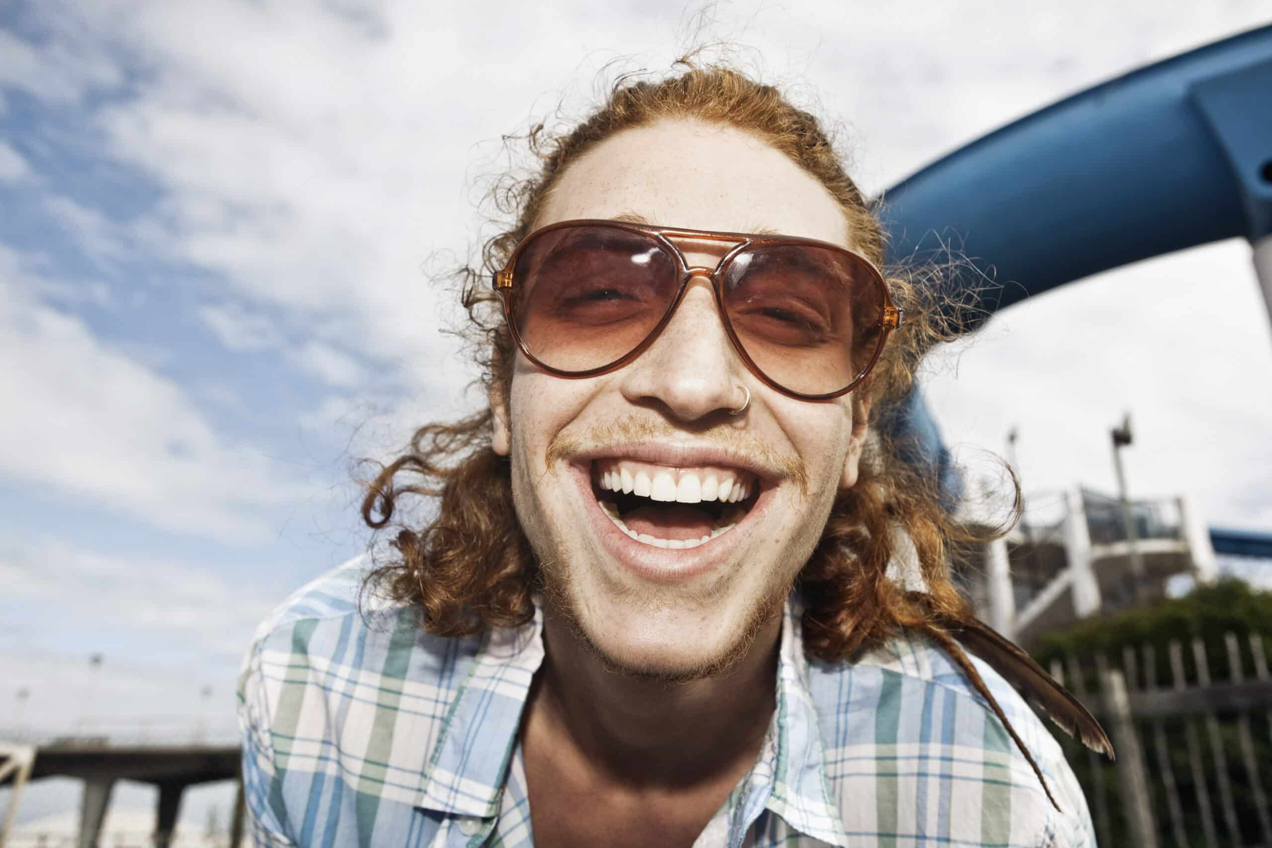 Eine Person mit langen lockigen Haaren und Sonnenbrille lächelt breit. Sie trägt ein kariertes Hemd. Im Hintergrund sind eine blaue Struktur und ein teilweise bewölkter Himmel zu sehen. © Fotografie Tomas Rodriguez