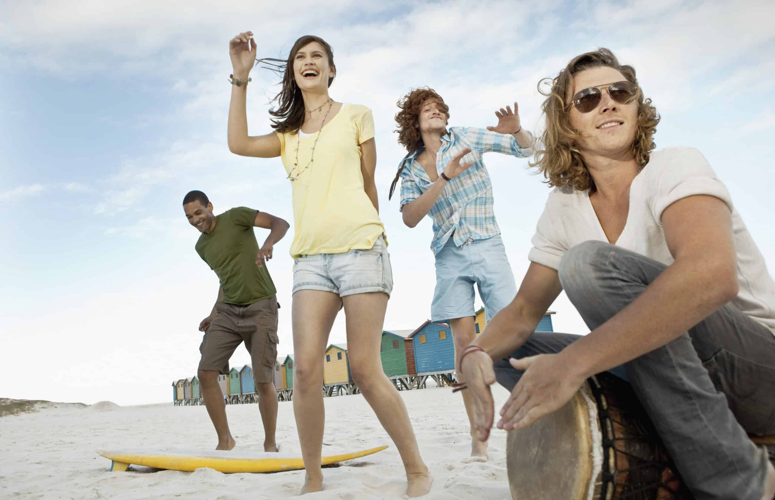 Vier junge Erwachsene genießen einen sonnigen Tag am Strand. Eine Person im grünen Hemd steht auf einem Surfbrett, eine andere in gelbem Oberteil und Jeansshorts tanzt, eine Person in blauem Karomuster reckt fröhlich die Arme in die Höhe und eine andere in Weiß spielt eine Trommel. Im Hintergrund stehen bunte Hütten. © Fotografie Tomas Rodriguez