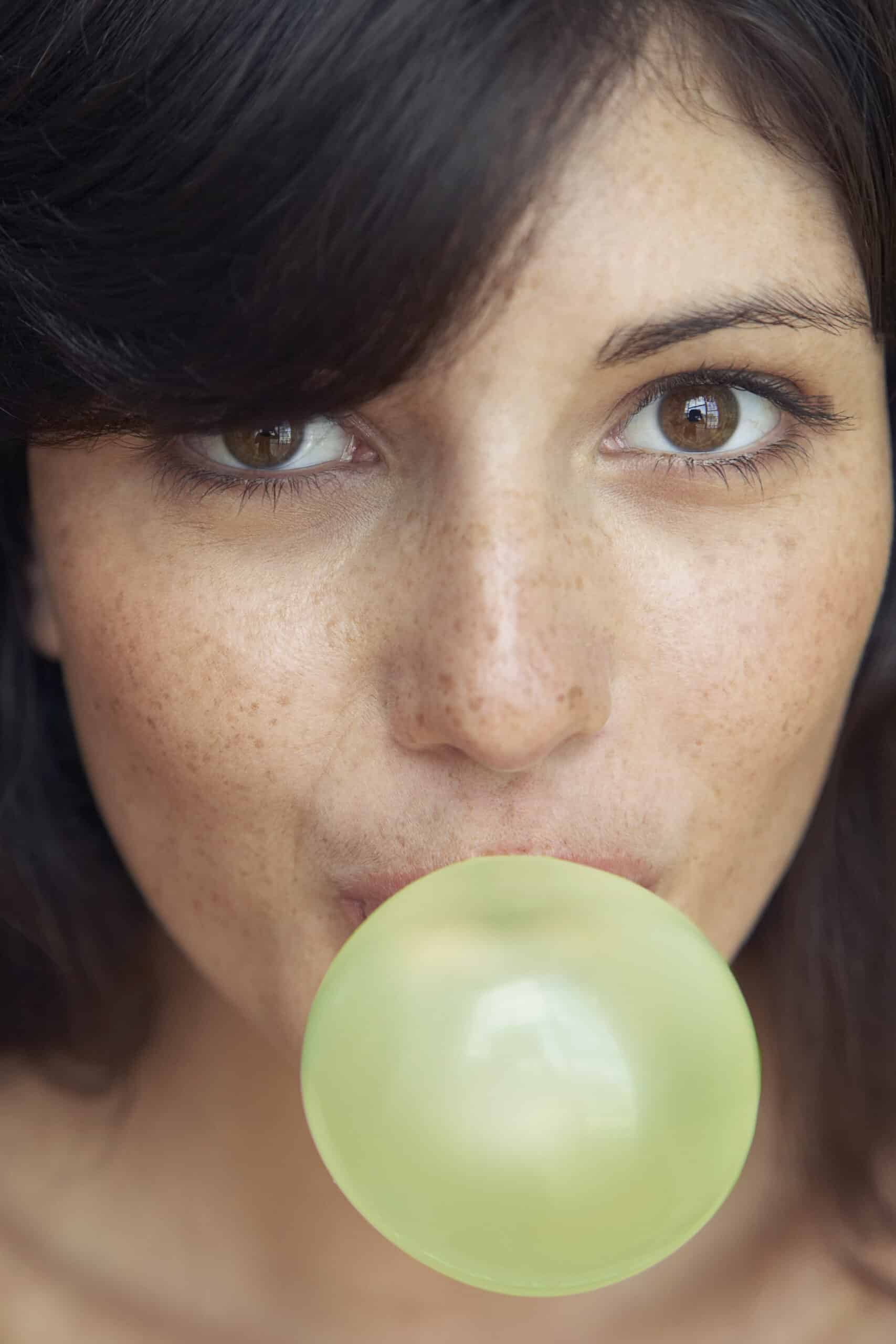 Eine Nahaufnahme einer Person mit dunklem Haar und braunen Augen, die eine große Kaugummiblase macht. Die Person hat sichtbare Sommersprossen und leicht geöffnete Lippen um die Blase herum. Der Hintergrund ist nicht klar zu erkennen, der Fokus liegt vollständig auf dem Gesicht der Person und der Blase. © Fotografie Tomas Rodriguez