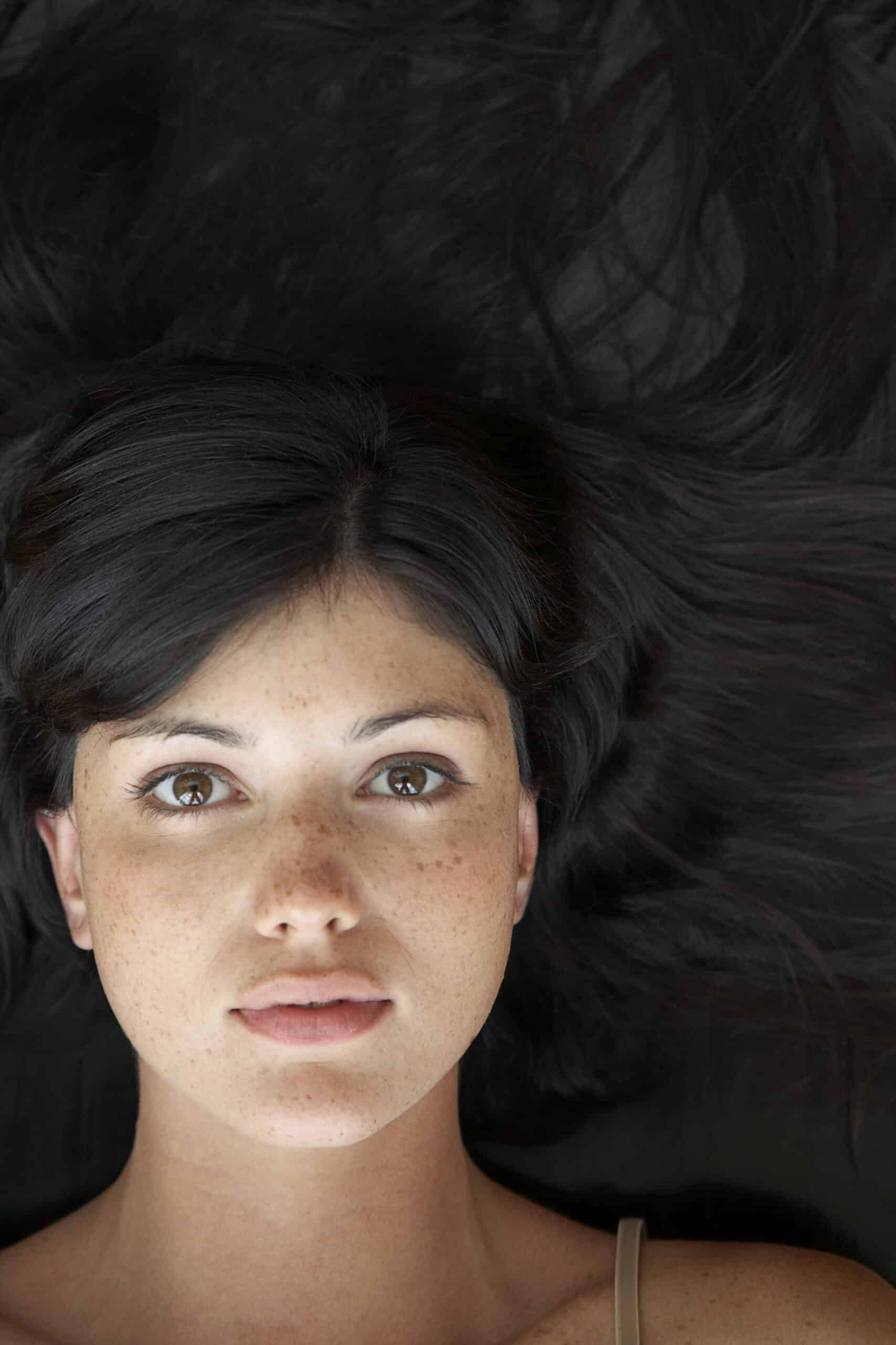 Eine Frau mit dunklem Haar liegt da, ihr Haar ist um ihren Kopf herum auf einer dunklen Oberfläche ausgebreitet. Sie hat einen neutralen Gesichtsausdruck, ihre Augen sind weit geöffnet und sie blickt direkt in die Kamera. Sie trägt ein dünnes Trägertop. © Fotografie Tomas Rodriguez