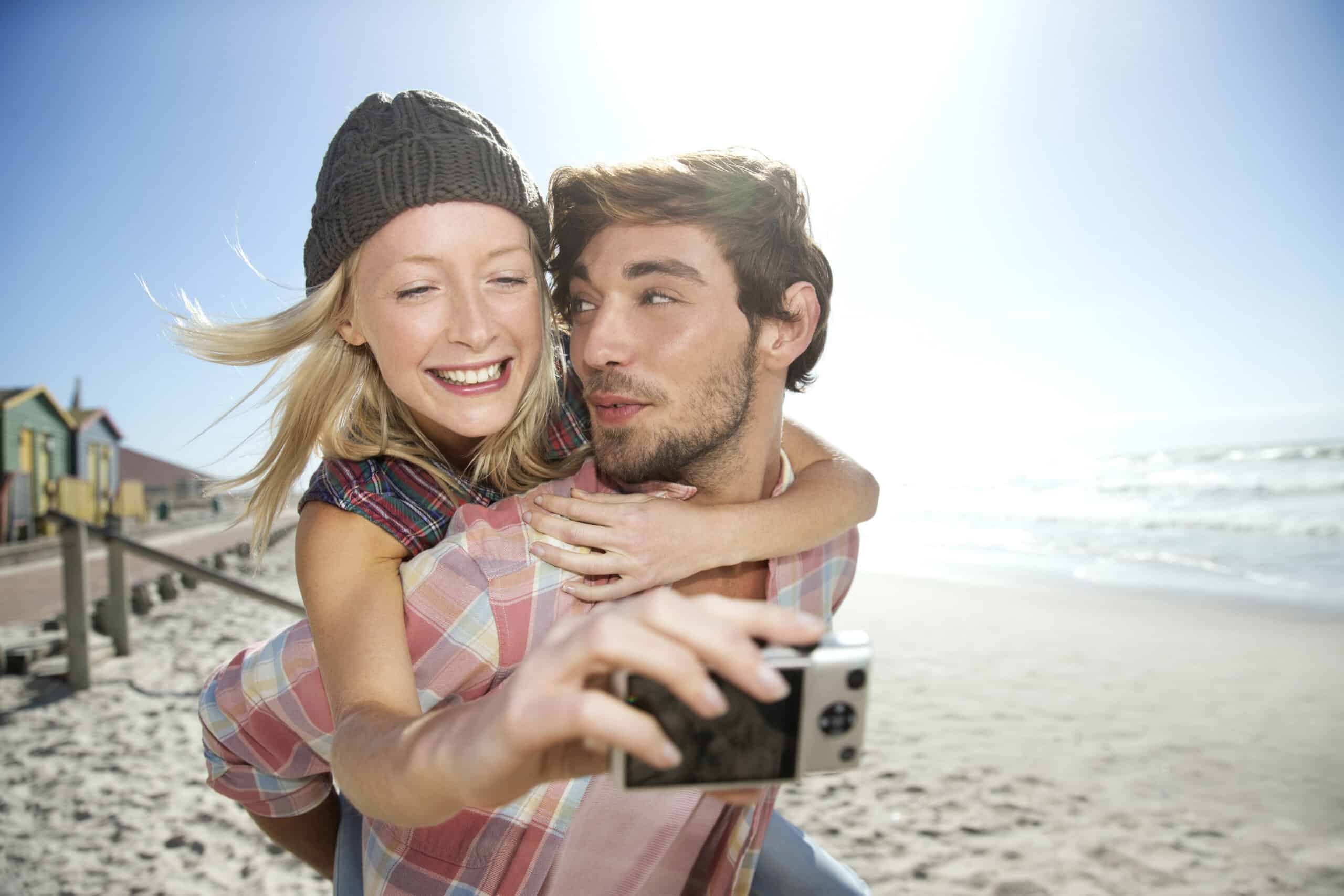 Eine lächelnde Frau mit einer Beanie sitzt auf dem Rücken eines Mannes und macht an einem sonnigen Strand ein Selfie. Im Hintergrund sind bunte Strandhütten und Meereswellen zu sehen. Beide sind leger gekleidet und scheinen einen spielerischen Moment zu genießen. © Fotografie Tomas Rodriguez