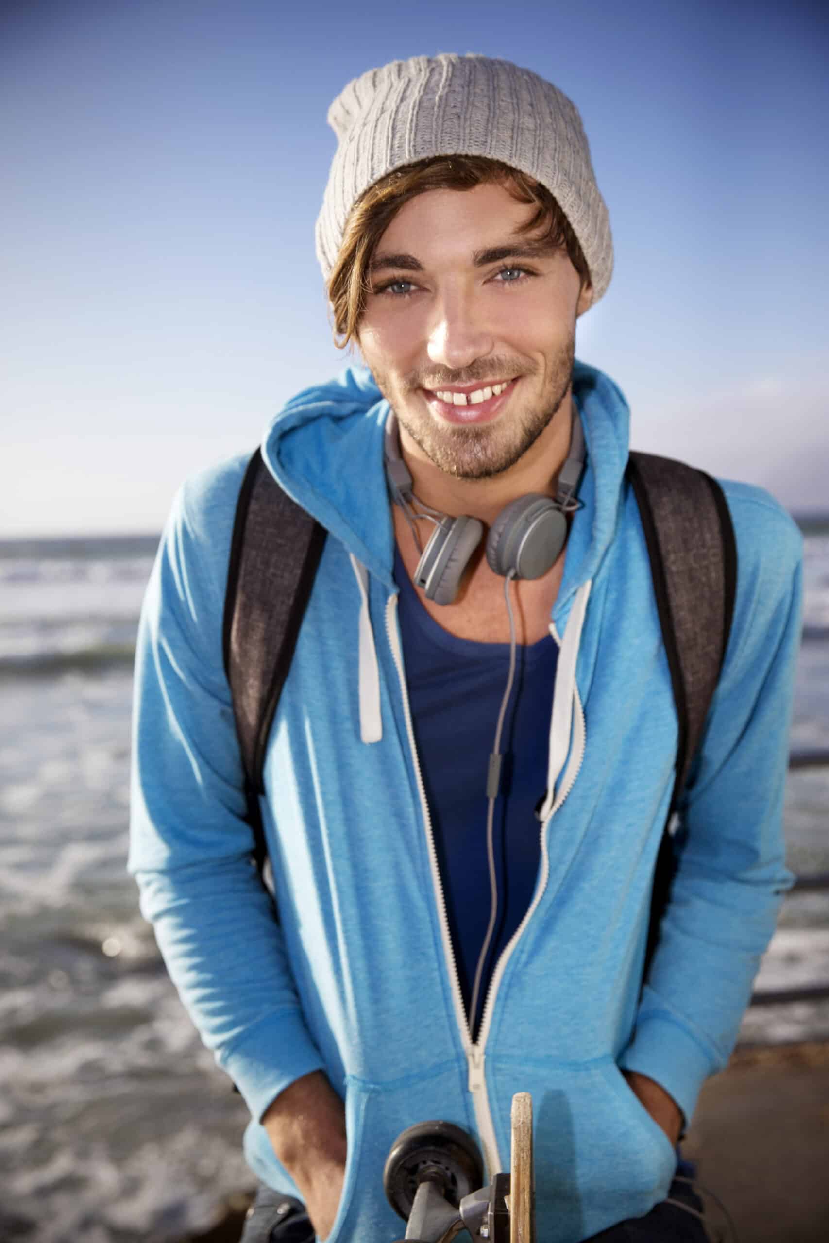 Ein junger Mann mit blauen Augen und einem leichten Lächeln steht am Meer. Er trägt eine graue Mütze, einen blauen Kapuzenpullover, ein marineblaues Hemd, einen Rucksack und Kopfhörer um den Hals. Er hat die Hände in den Taschen und ein Skateboard vor sich. Der Himmel ist klar und im Hintergrund sind die Wellen zu sehen. © Fotografie Tomas Rodriguez