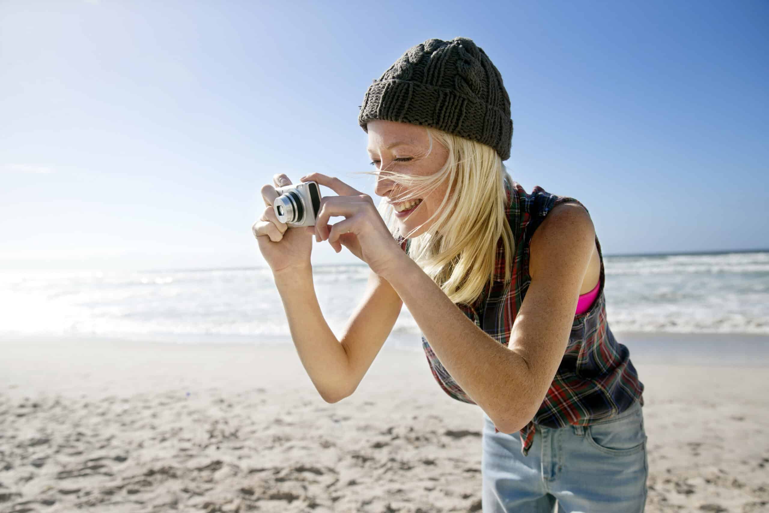 Eine Frau steht an einem Sandstrand, hält eine Kamera in der Hand und lächelt, während sie ein Foto macht. Sie trägt eine graue Strickmütze, ein ärmelloses kariertes Hemd und Jeans. Im Hintergrund sind die Meereswellen und ein klarer blauer Himmel zu sehen. © Fotografie Tomas Rodriguez