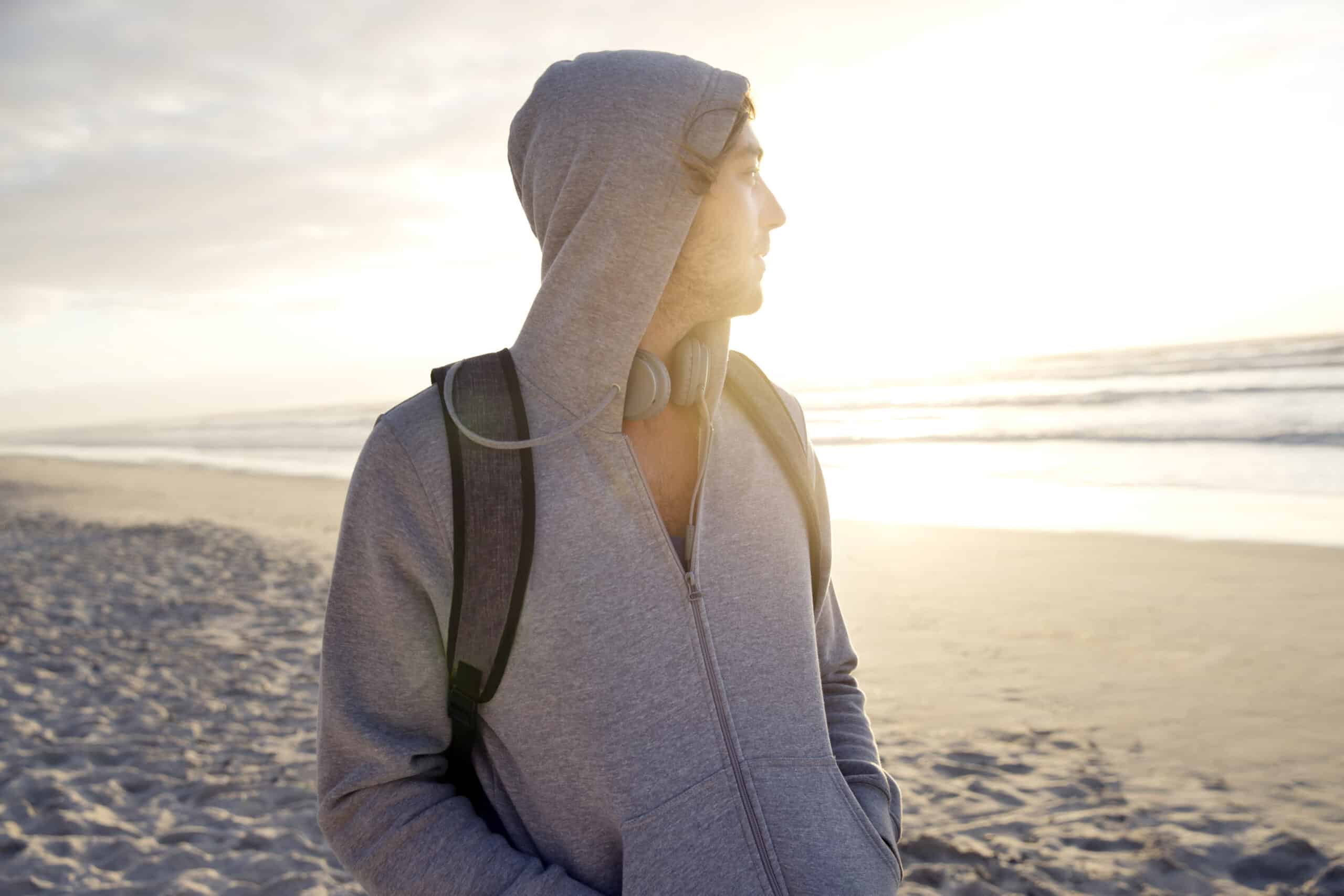 Eine Person in einem grauen Kapuzenpullover steht mit einem Rucksack und Kopfhörern um den Hals an einem Strand und blickt bei Sonnenuntergang auf das Meer. Der Himmel ist teilweise bewölkt und im Vordergrund ist der Sand zu sehen. © Fotografie Tomas Rodriguez