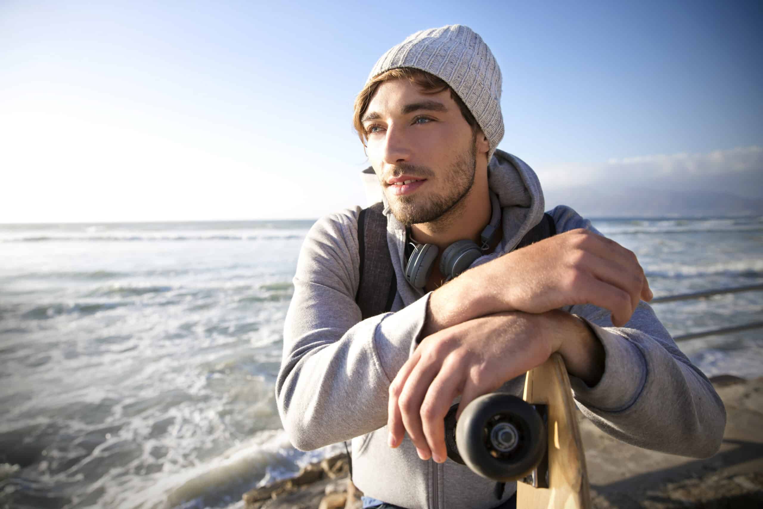 Ein Mann mit Beanie und Kapuzenpullover sowie Kopfhörern um den Hals lehnt auf einem Skateboard und blickt aufs Meer. Der Himmel ist klar und im Hintergrund brechen die Wellen sanft gegen das Ufer. © Fotografie Tomas Rodriguez
