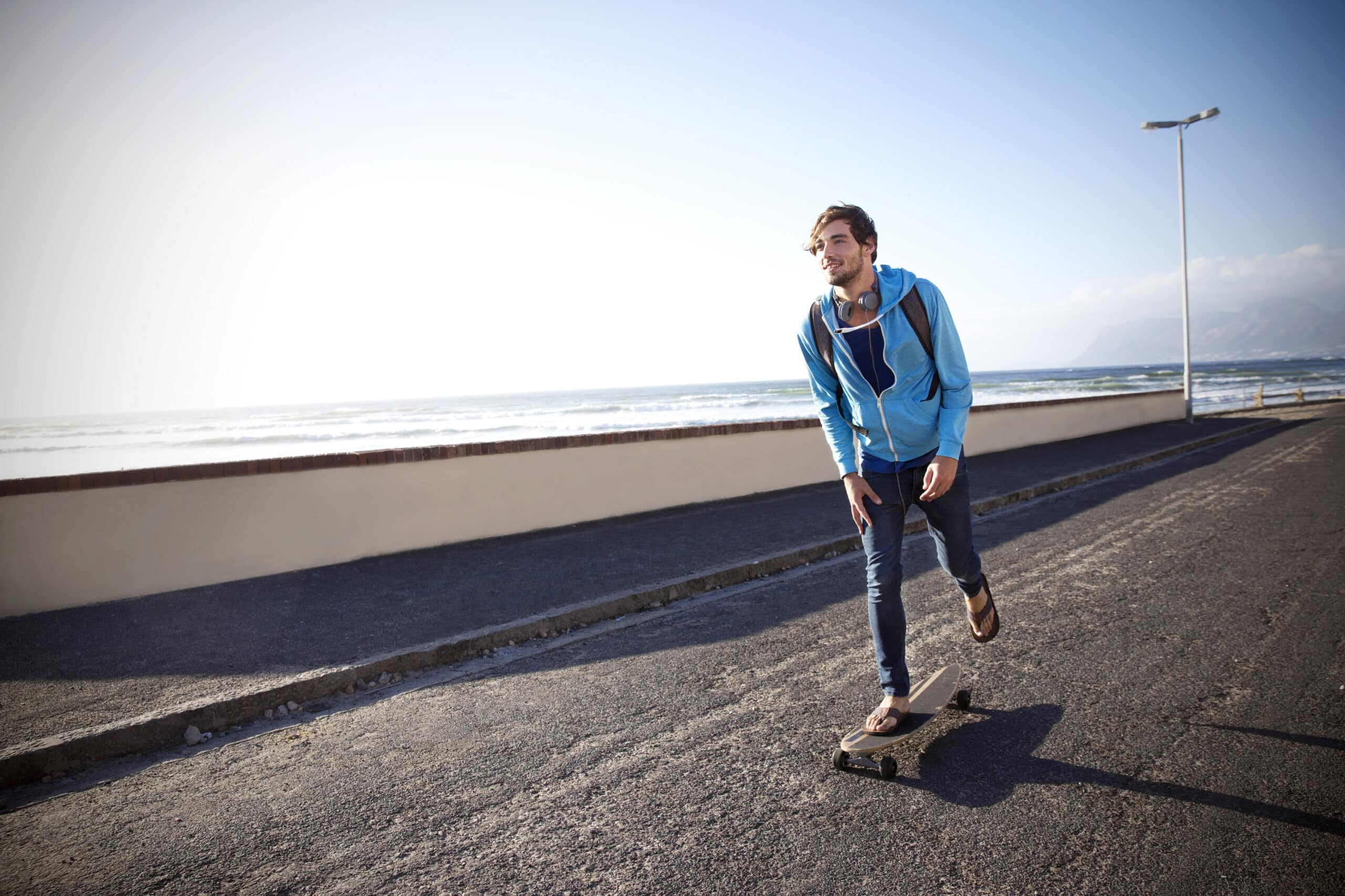 Eine Person in einer blauen Jacke und Jeans fährt auf einem gepflasterten Weg am Meer entlang Skateboard. Sie trägt Kopfhörer und einen Rucksack und blickt nach vorne, während sie an einem sonnigen Tag mit klarem Himmel skatet. Im Hintergrund sind der Meerblick und die Berge zu sehen. © Fotografie Tomas Rodriguez