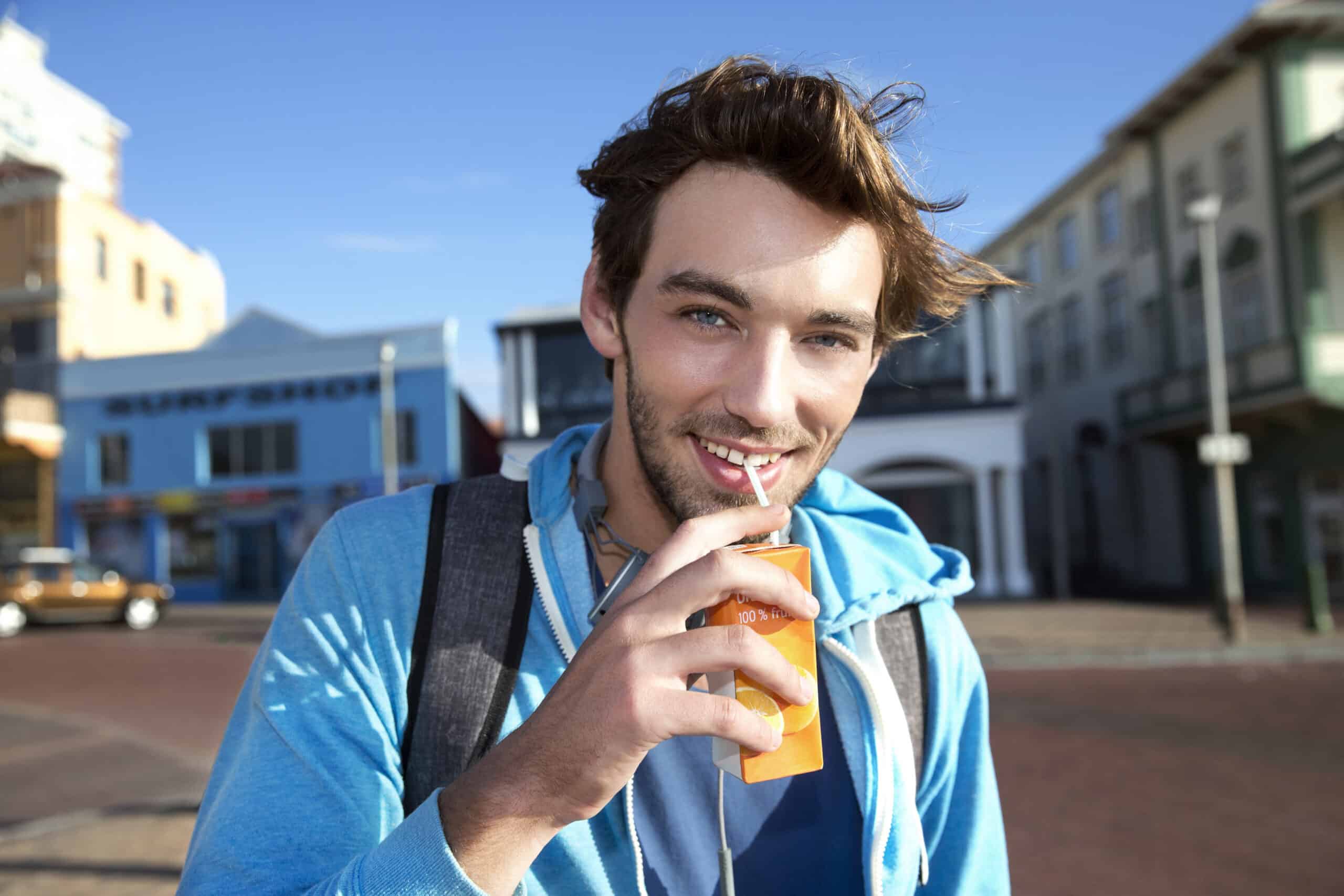Ein junger Mann mit zerzaustem braunem Haar trinkt mit einem Strohhalm aus einem Orangensaftkarton. Er trägt einen blauen Kapuzenpullover und einen Rucksack. Im Hintergrund sind ein klarer blauer Himmel und farbenfrohe Gebäude entlang einer Straße zu sehen. © Fotografie Tomas Rodriguez