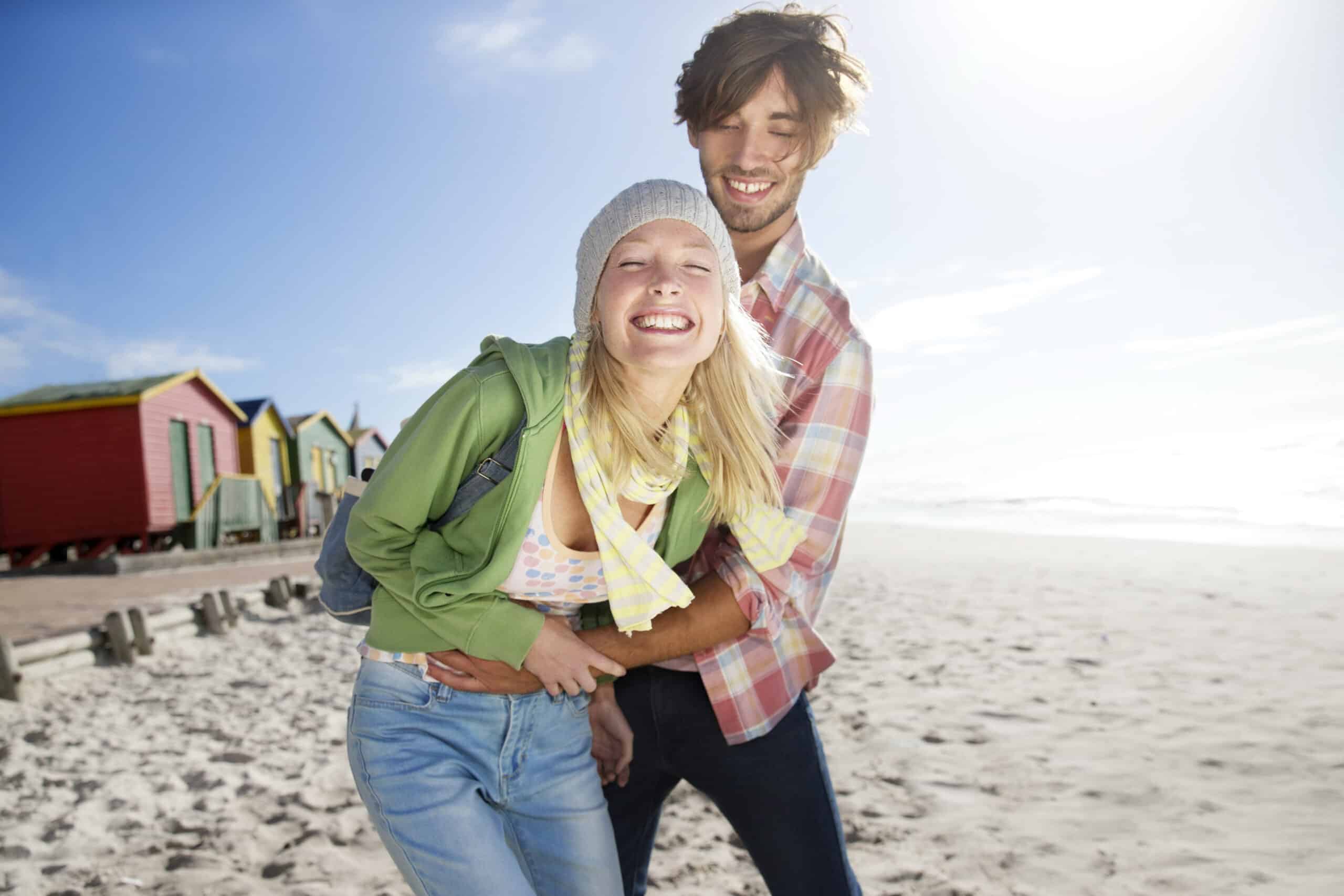Ein fröhlicher Mann und eine fröhliche Frau lachen zusammen an einem sonnigen Strand und stehen neben bunten Strandhütten. Der Mann hat seine Arme von hinten um die Frau gelegt. Der Sand erstreckt sich bis zum Meer unter einem strahlend blauen Himmel mit ein paar Wolken. © Fotografie Tomas Rodriguez