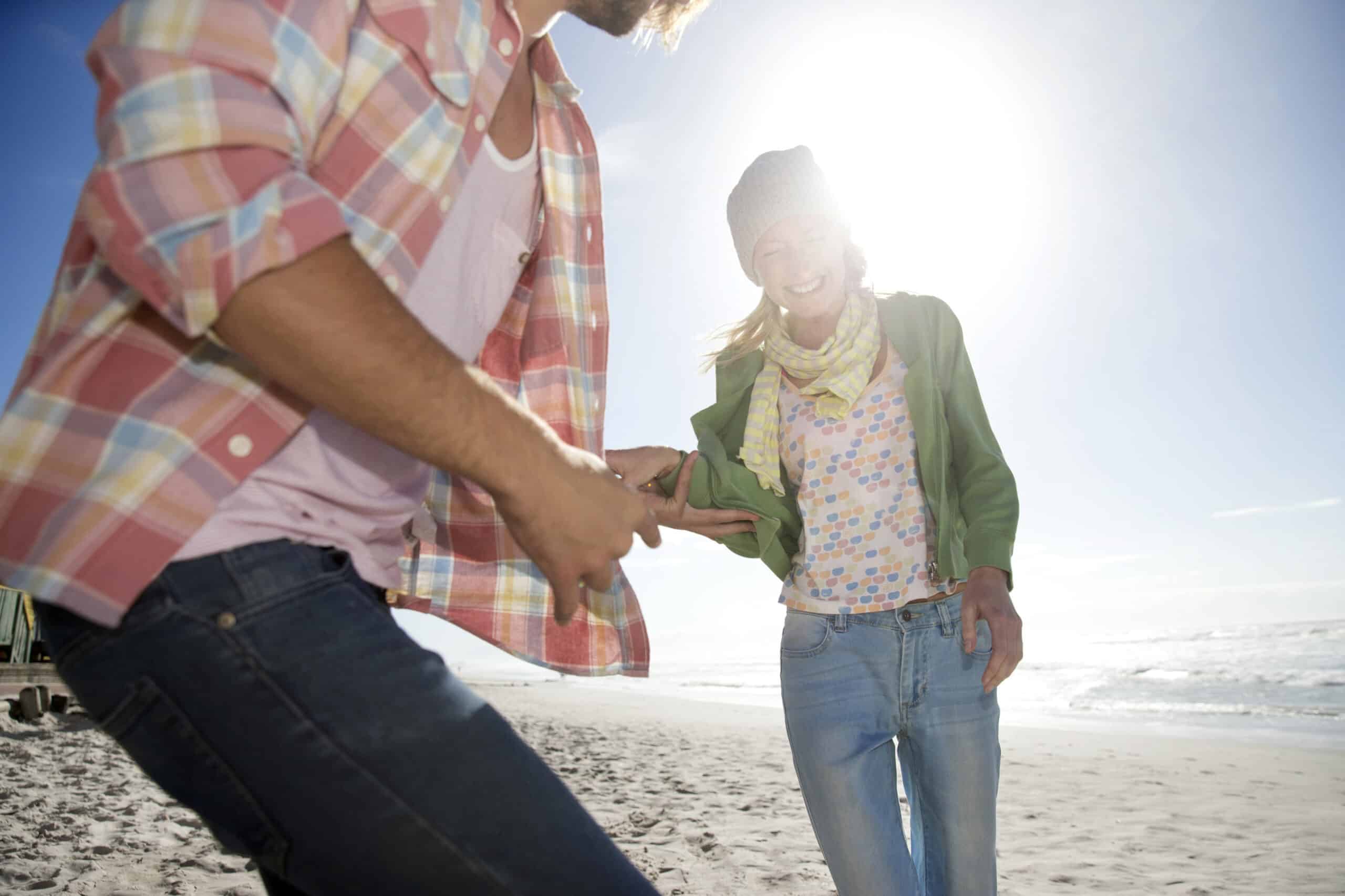 Zwei Menschen gehen an einem sonnigen Strand spazieren, halten Händchen und lächeln. Die Person im Vordergrund trägt ein kariertes Hemd, während die Person im Hintergrund eine Beanie-Mütze und eine grüne Jacke trägt. Die Sonne scheint hell und der Himmel ist klar. © Fotografie Tomas Rodriguez