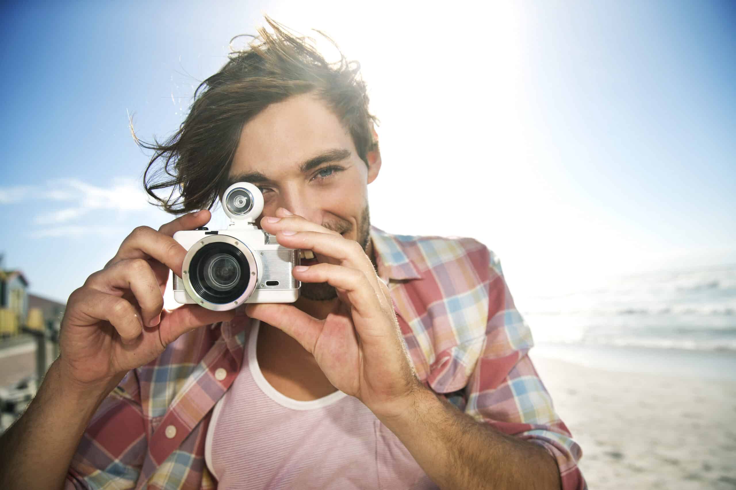 Eine Person mit schulterlangem Haar und kariertem Hemd steht an einem Strand und hält eine Kamera vor ihr Gesicht. Im Hintergrund scheint hell die Sonne und verleiht der Szene eine warme, sonnige Atmosphäre. © Fotografie Tomas Rodriguez