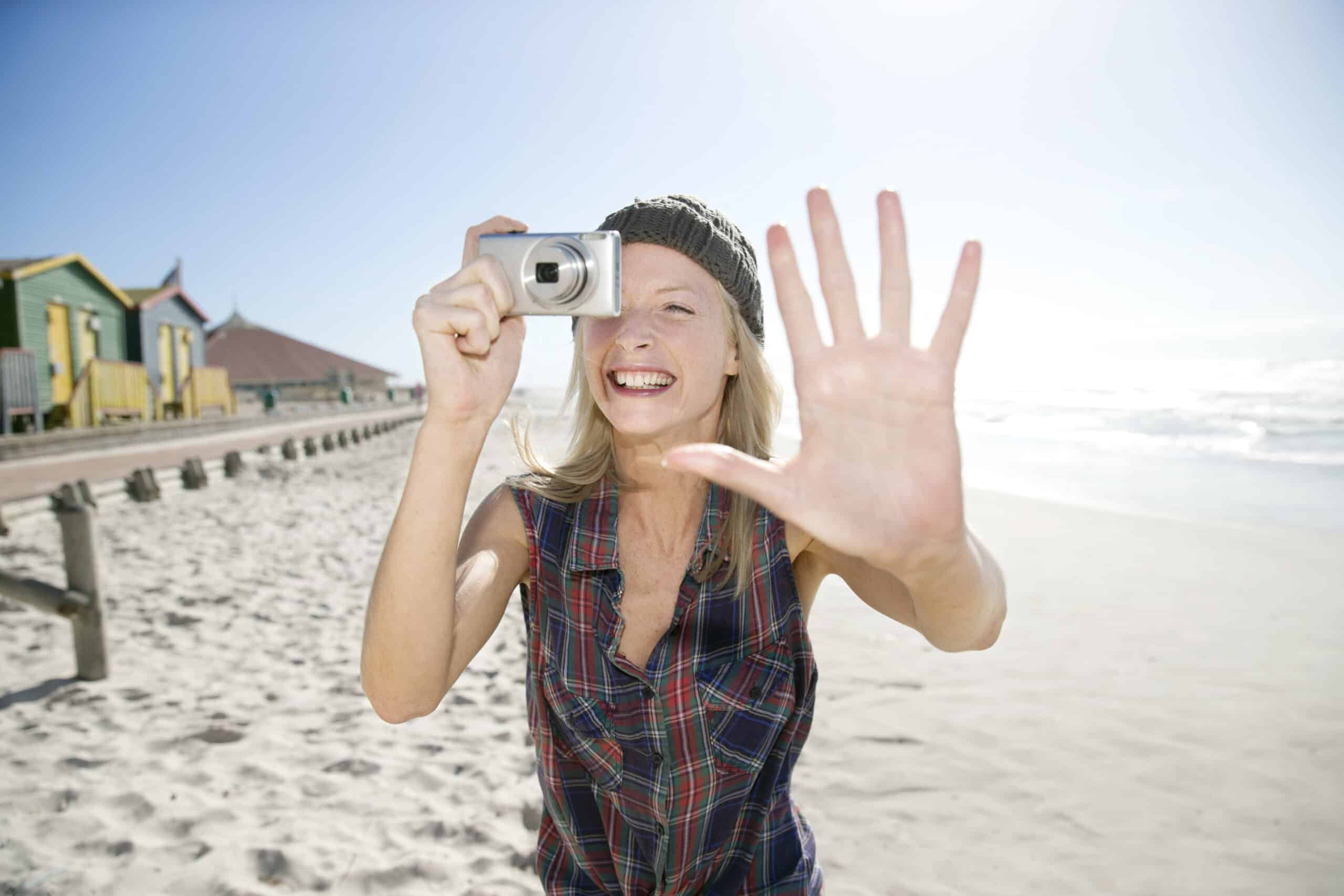 Eine Person mit langen Haaren, einer Beanie-Mütze und einem karierten Hemd steht an einem Strand, macht ein Foto und lächelt breit. Ihre andere Hand ist erhoben und blickt in die Kamera. Im Hintergrund entlang der Küste sind bunte Strandhütten zu sehen. © Fotografie Tomas Rodriguez