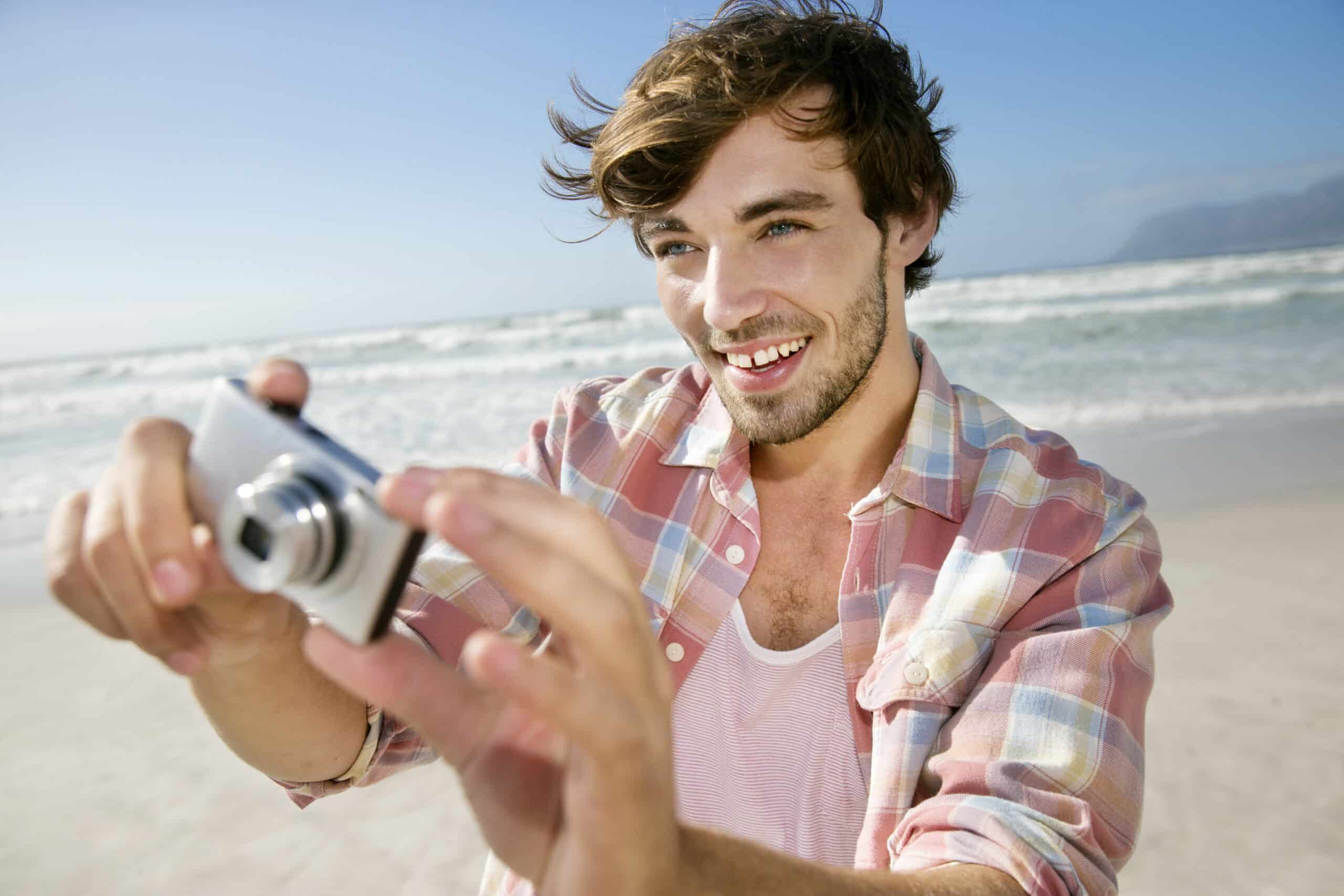Ein Mann mit Bart und zerzaustem Haar lächelt, während er mit einer Digitalkamera ein Foto macht. Er trägt ein rosa-weiß kariertes Hemd über einem weißen Tanktop. Im Hintergrund ist ein sonniger Strand mit Wellen und einem klaren blauen Himmel zu sehen. © Fotografie Tomas Rodriguez