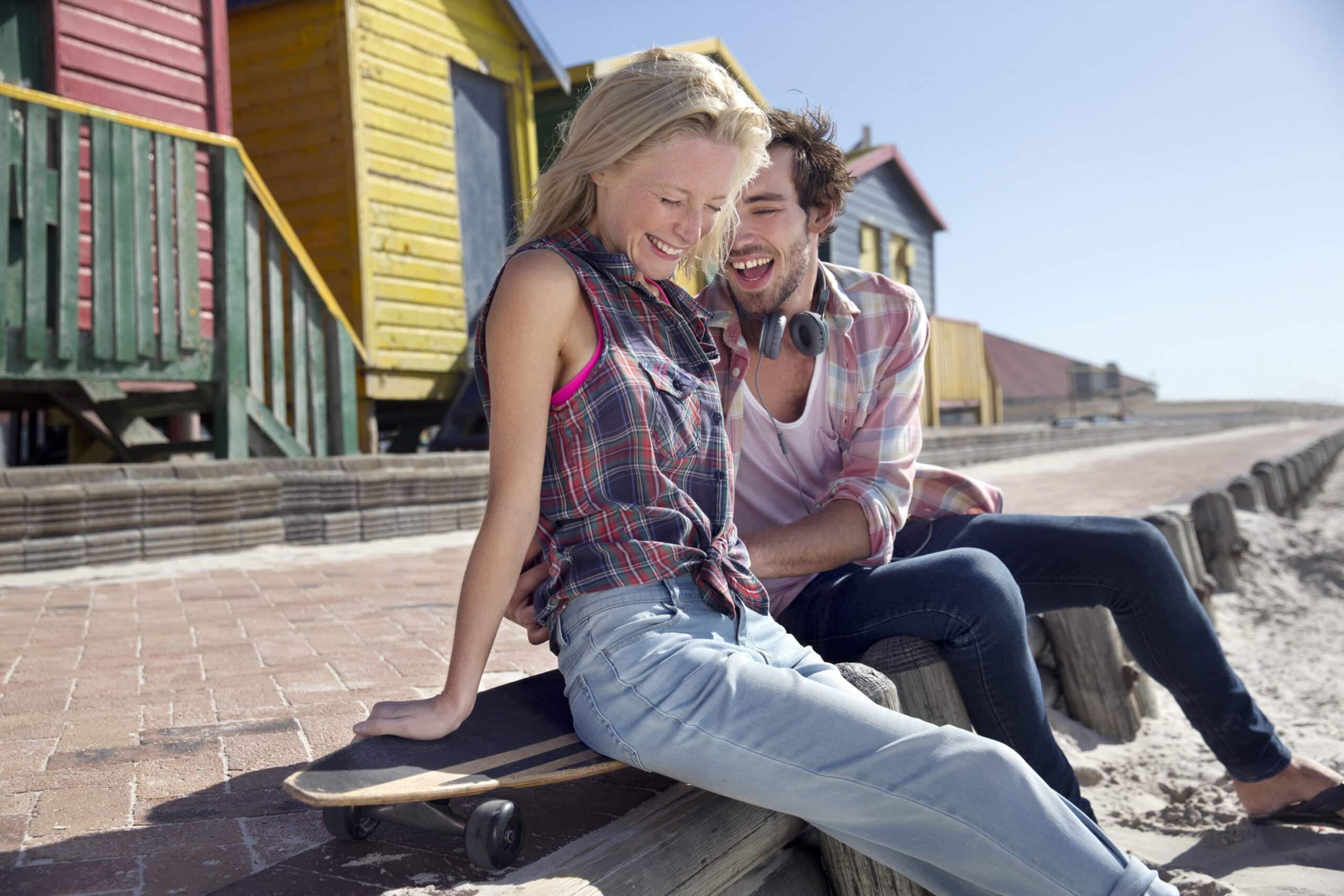 Eine Frau und ein Mann lachen, während sie auf einem Holzbalken am Strand sitzen. Die Frau sitzt in einem karierten Hemd auf einem Skateboard und der Mann steht mit Kopfhörern um den Hals neben ihr. Im Hintergrund sind bunte Strandhütten unter einem klaren Himmel zu sehen. © Fotografie Tomas Rodriguez