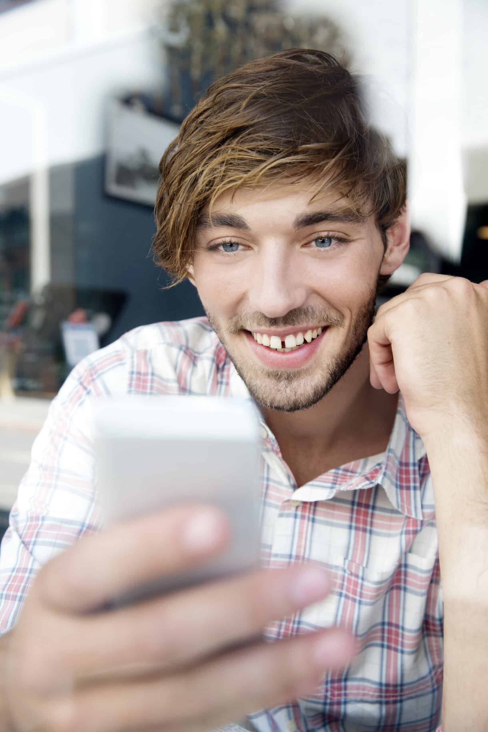 Ein junger Mann mit hellbraunem Haar und Bart lächelt, während er auf sein Smartphone schaut. Er trägt ein kariertes Hemd und scheint sich in einem hellen Innenraum zu befinden, möglicherweise in einem Café. Er stützt seinen Kopf auf eine Hand und scheint mit dem beschäftigt zu sein, was er auf seinem Telefon ansieht. © Fotografie Tomas Rodriguez