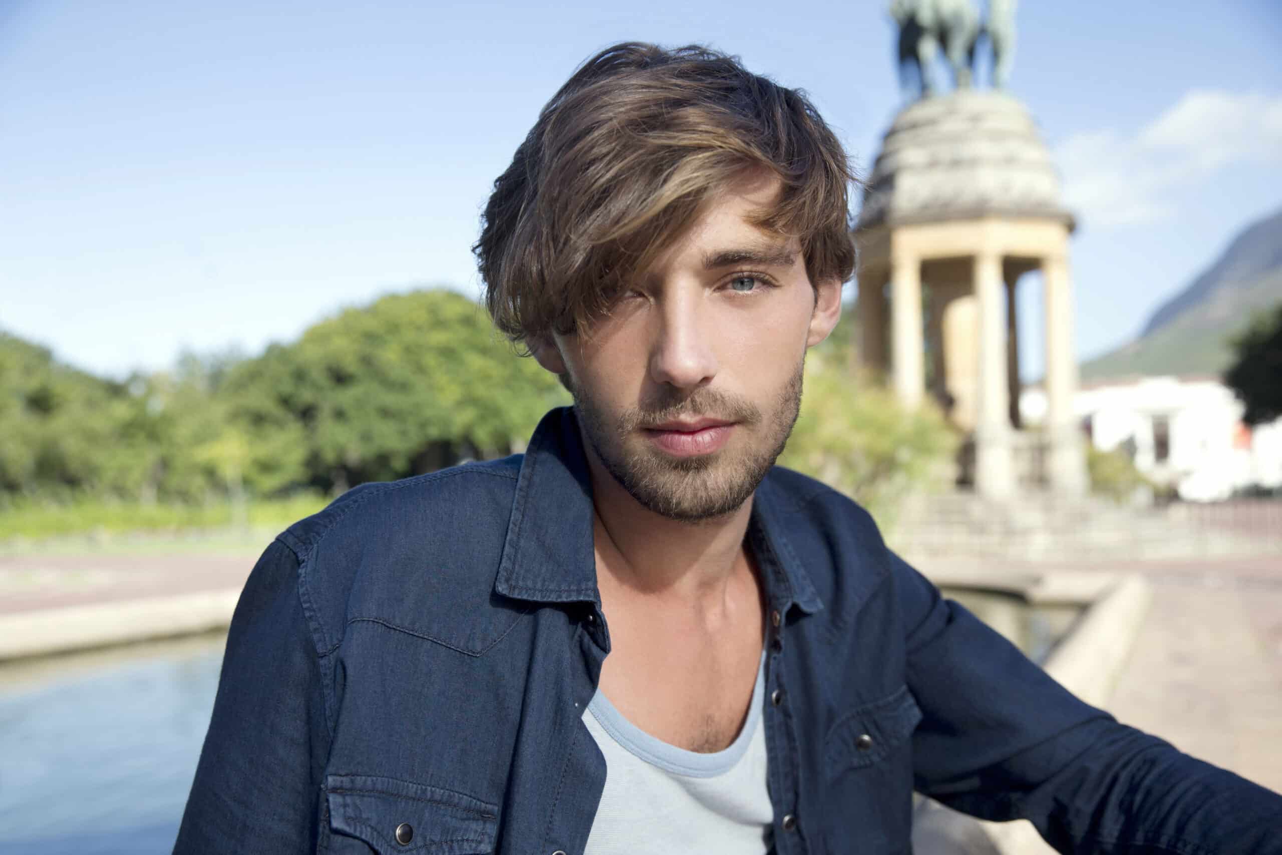 Ein junger Mann mit zerzaustem Haar, der ein Jeanshemd über einem hellgrauen T-Shirt trägt, steht vor einem Brunnen. Im Hintergrund ist ein Steindenkmal mit einer Statue darauf zu sehen, und üppiges Grün umgibt die Gegend. Der Himmel ist klar und blau. © Fotografie Tomas Rodriguez