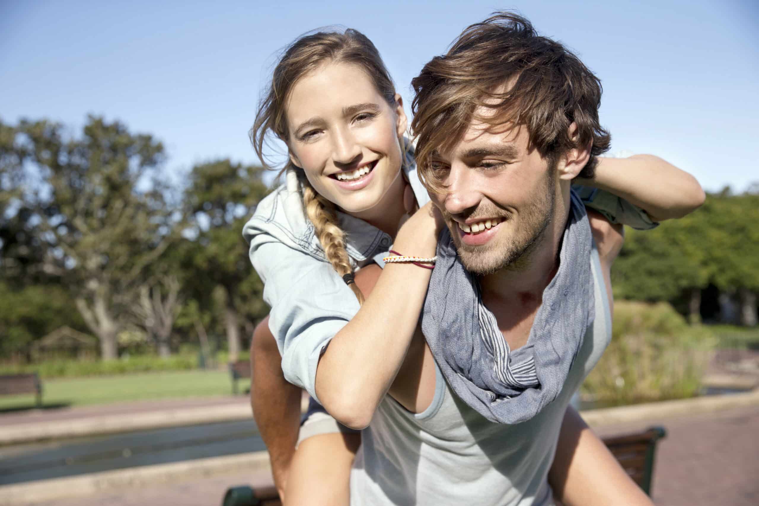 Eine junge Frau mit Zopf und ein junger Mann mit zerzaustem Haar lächeln an einem sonnigen Tag in einem Park. Der Mann trägt die Frau Huckepack. Im Hintergrund sind Bäume und ein Weg zu sehen. © Fotografie Tomas Rodriguez