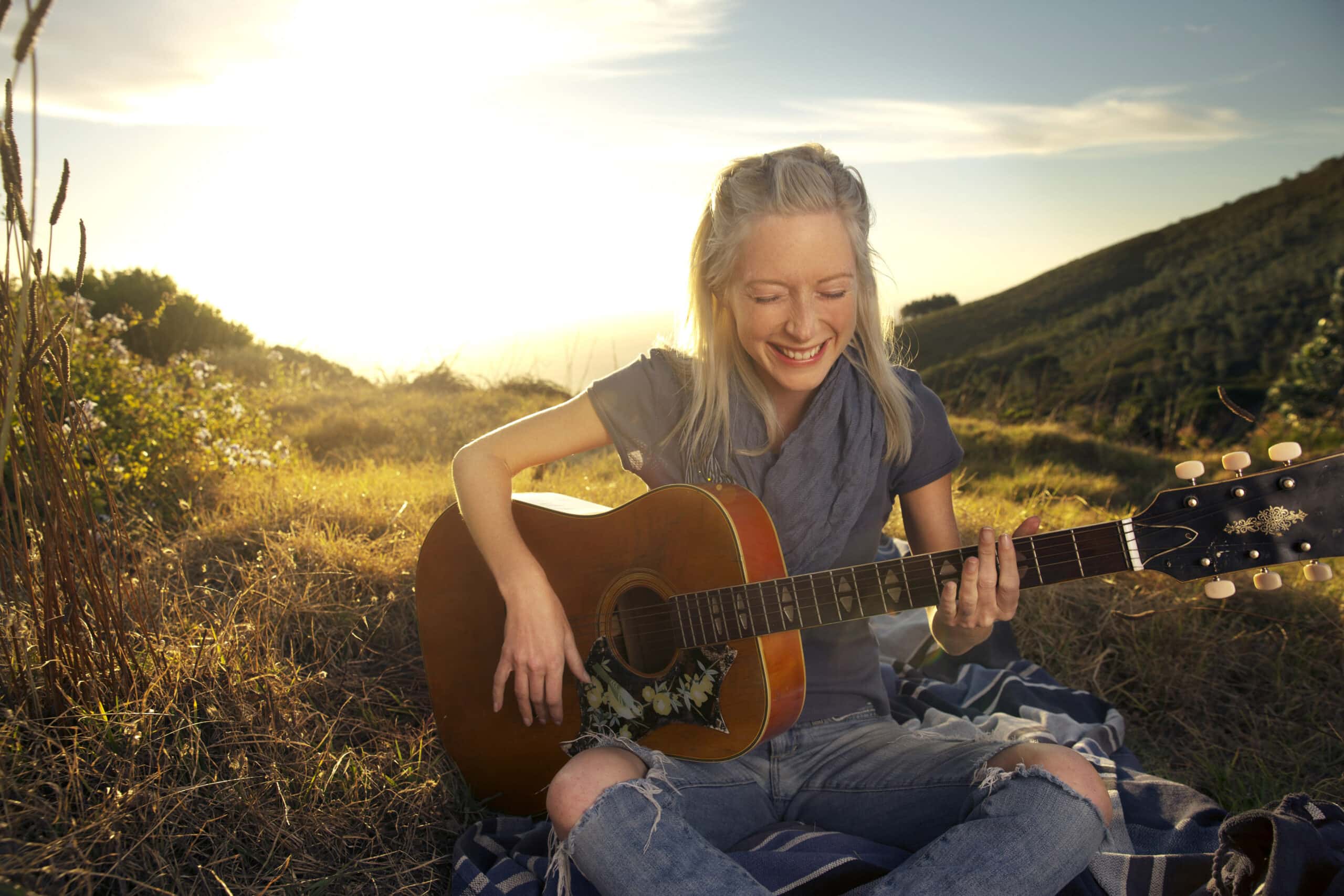 Eine lächelnde Frau mit langen blonden Haaren sitzt auf einer Decke auf einer Wiese und spielt bei Sonnenuntergang Akustikgitarre. Sie trägt ein graues Hemd und zerrissene Jeans. Im Hintergrund sind Hügel und ein klarer Himmel zu sehen, was eine heitere und fröhliche Atmosphäre schafft. © Fotografie Tomas Rodriguez