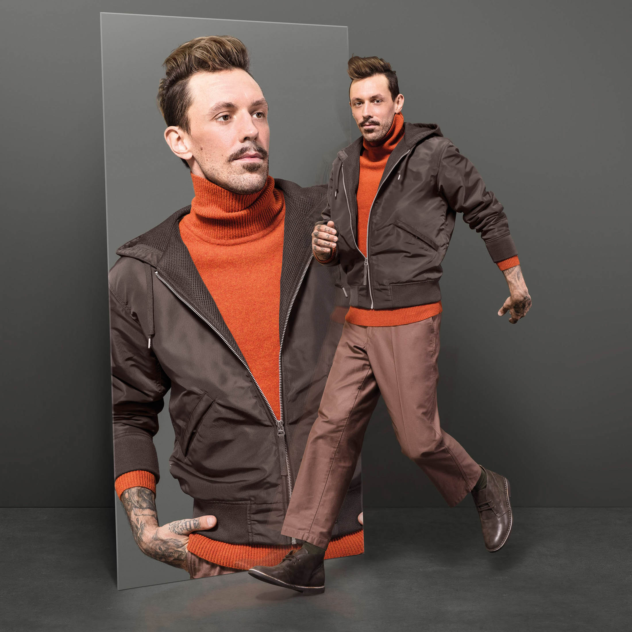 Ein Mann in einer eleganten braunen Jacke, einem orangefarbenen Pullover und einer braunen Hose ist in zwei Posen zu sehen: eine in ruhiger Pose und eine andere mitten in der Bewegung, als würde er vor einem grauen Hintergrund rennen. © Fotografie Tomas Rodriguez