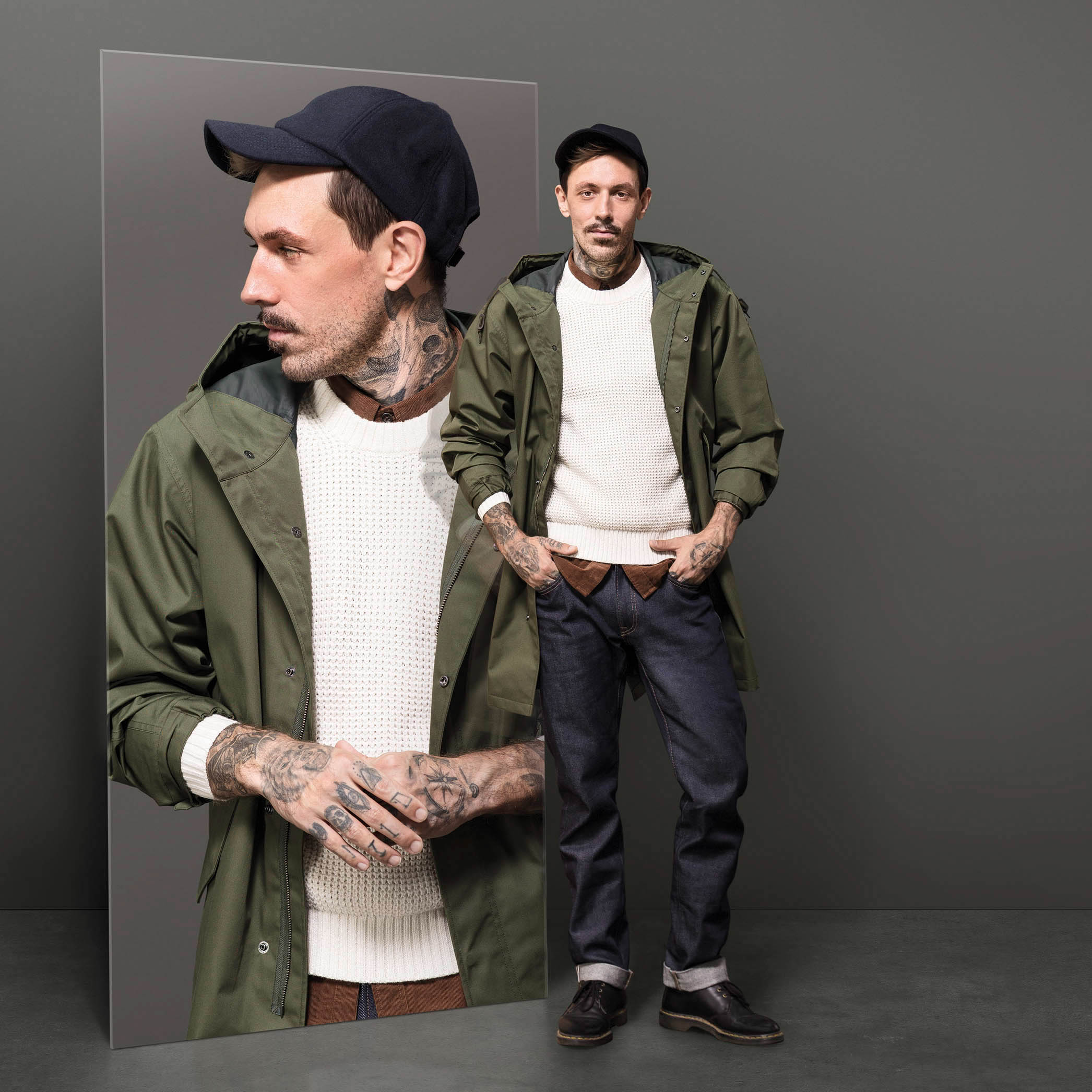 Ein Mann mit Tätowierungen steht selbstbewusst in einer grünen Jacke, einem weißen Pullover, Jeans und einer Mütze neben einem Spiegelbild seines Profils. © Fotografie Tomas Rodriguez