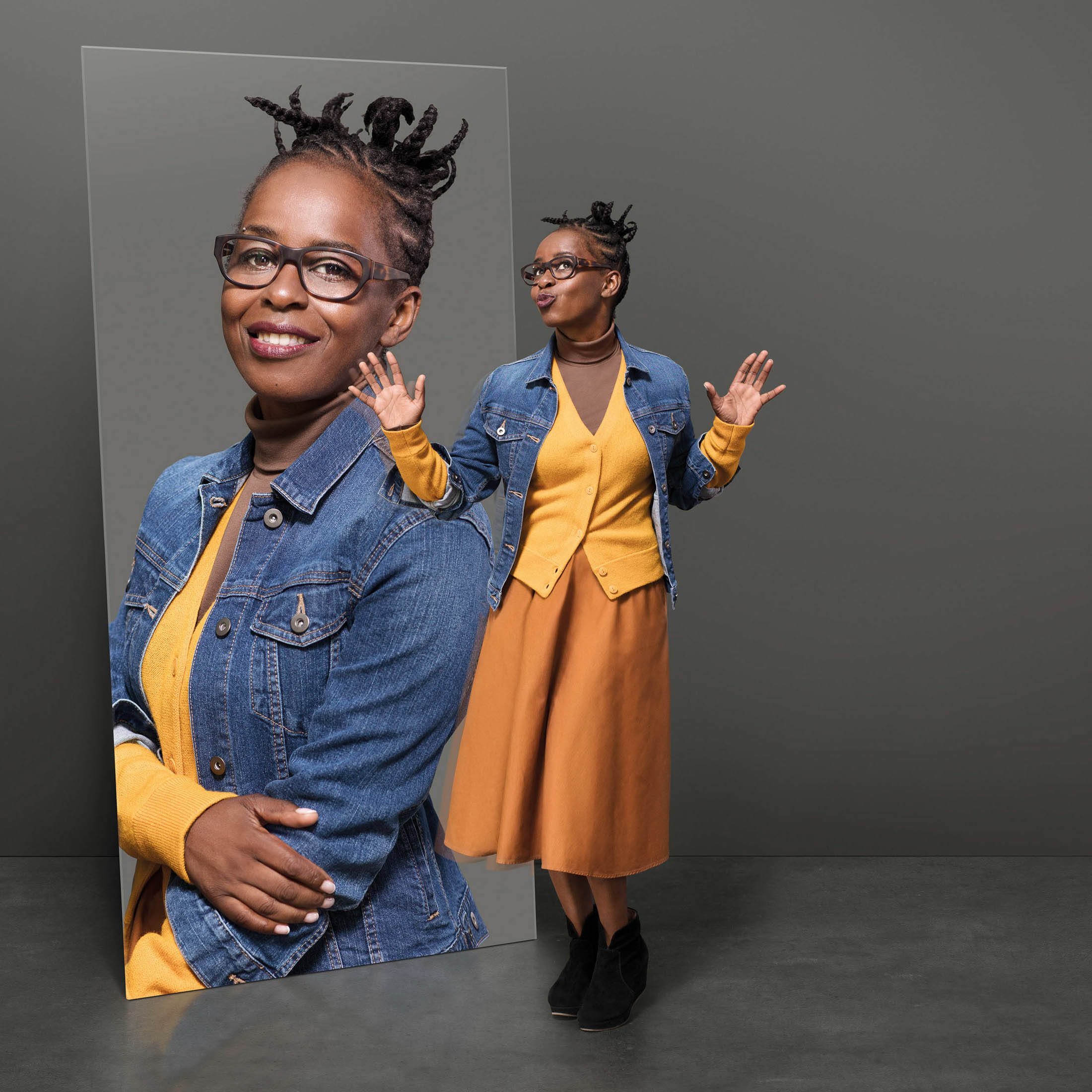 Eine lächelnde schwarze Frau mit Brille und gestyltem Haar ist in einem Doppelporträt zu sehen. Im Hintergrund posiert sie mit erhobenen Händen, während der Vordergrund auf ihr Gesicht fokussiert ist, alles vor einem grauen Hintergrund. © Fotografie Tomas Rodriguez