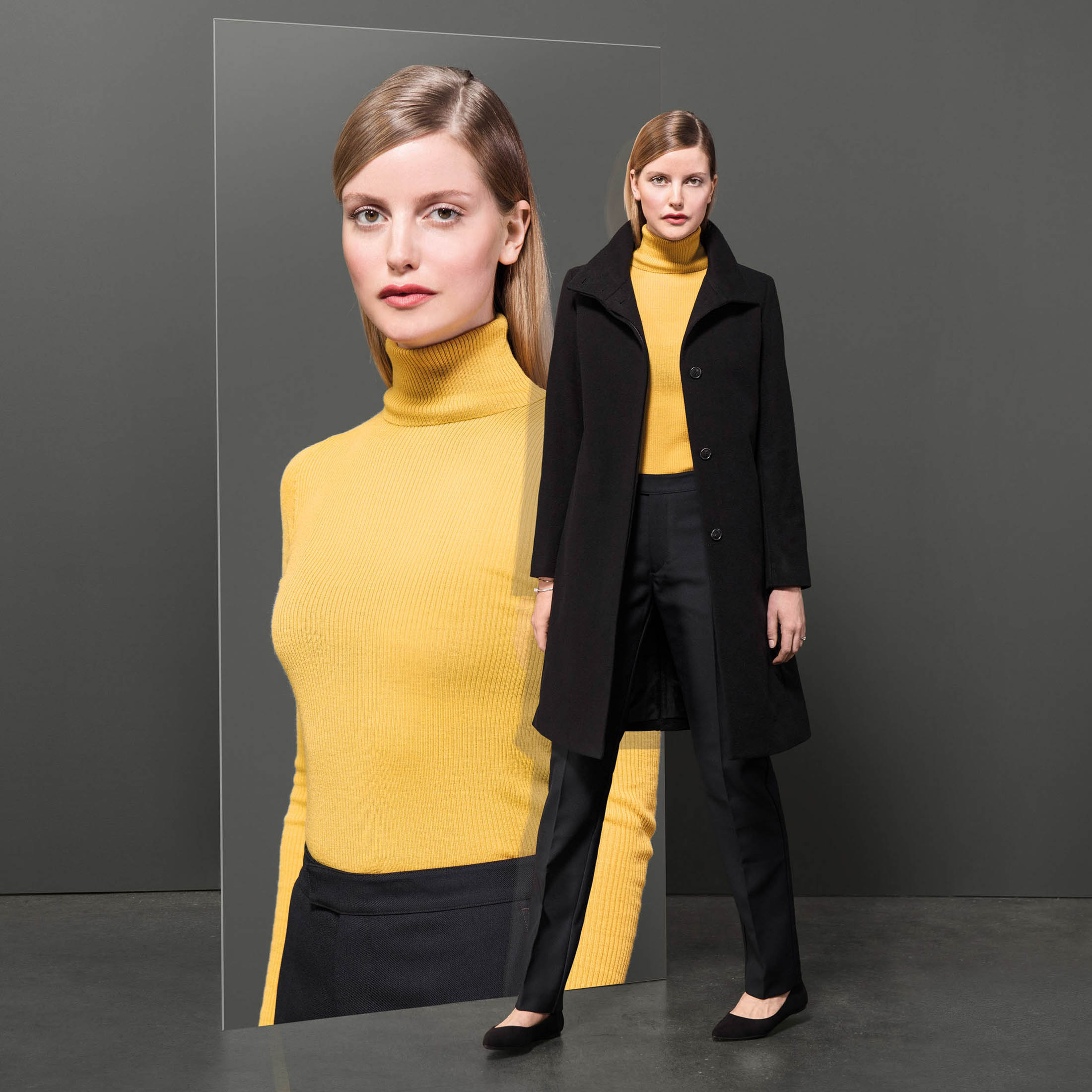 Eine Frau in einem modischen gelben Rollkragenpullover und schwarzen Hosen steht neben einem Spiegel, der ihr Bild reflektiert, und trägt über dem Rollkragenpullover einen schicken schwarzen Mantel. © Fotografie Tomas Rodriguez