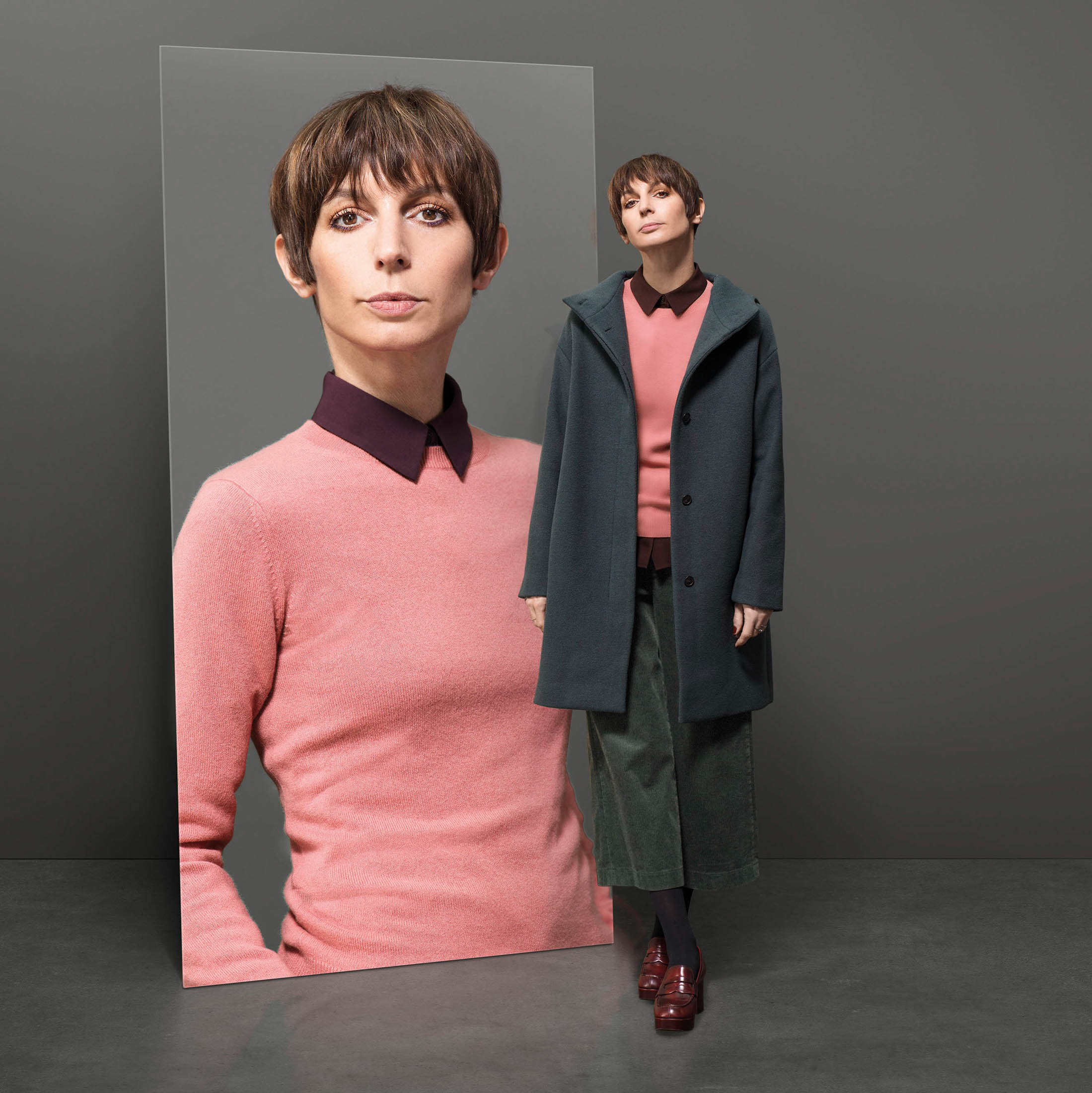 Eine Frau mit kurzen braunen Haaren ist in einem Studio abgebildet. Sie steht neben einem Spiegel, der sie in einem dunklen Mantel über einem rosa Pullover und einem grünen Rock vor einem grauen Hintergrund reflektiert. © Fotografie Tomas Rodriguez