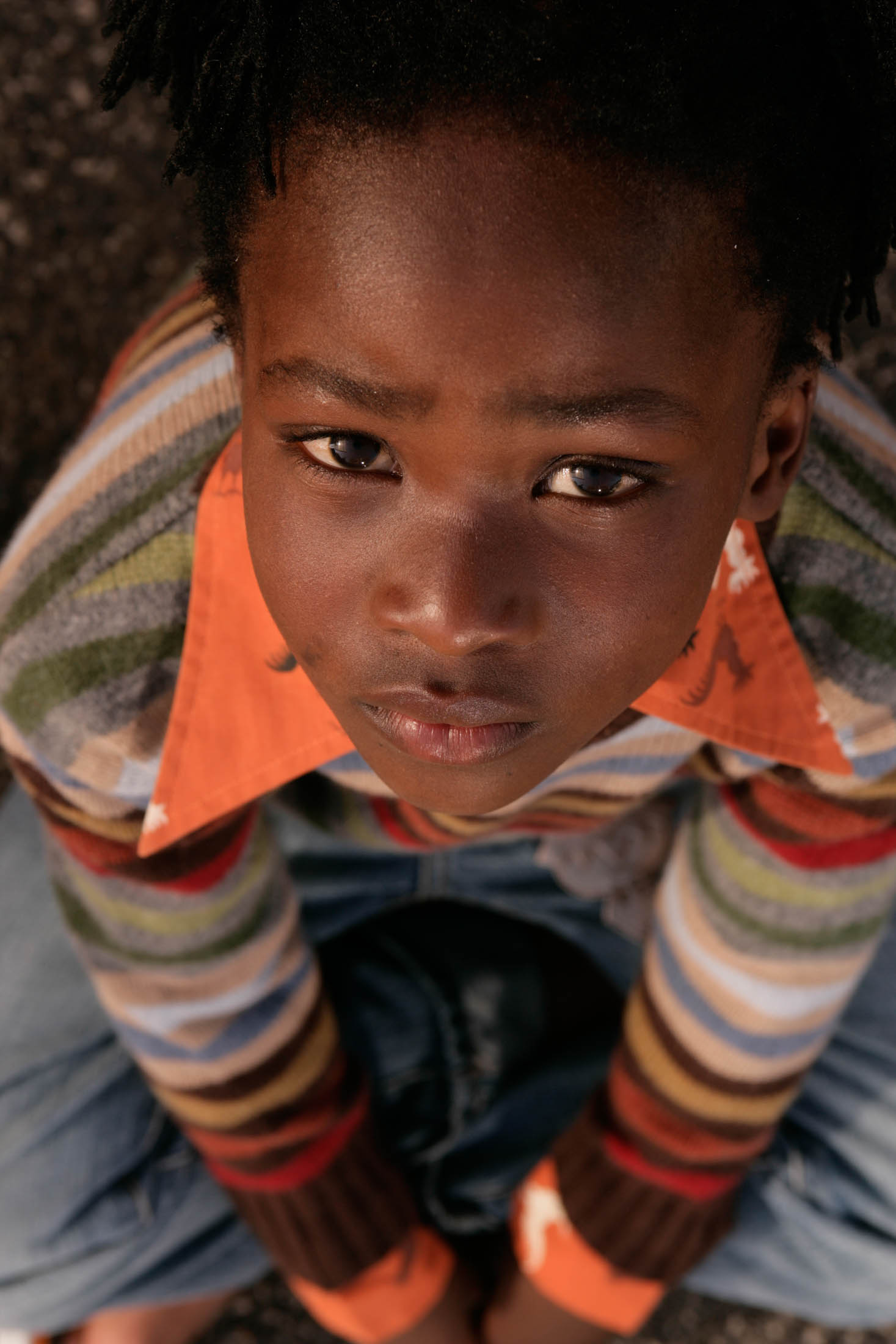 Ein Nahaufnahme-Porträt eines kleinen Jungen mit dunkler Haut und Dreadlocks, der direkt in die Kamera blickt. Er trägt einen bunt gestreiften Pullover und einen orangefarbenen Kragen. © Fotografie Tomas Rodriguez