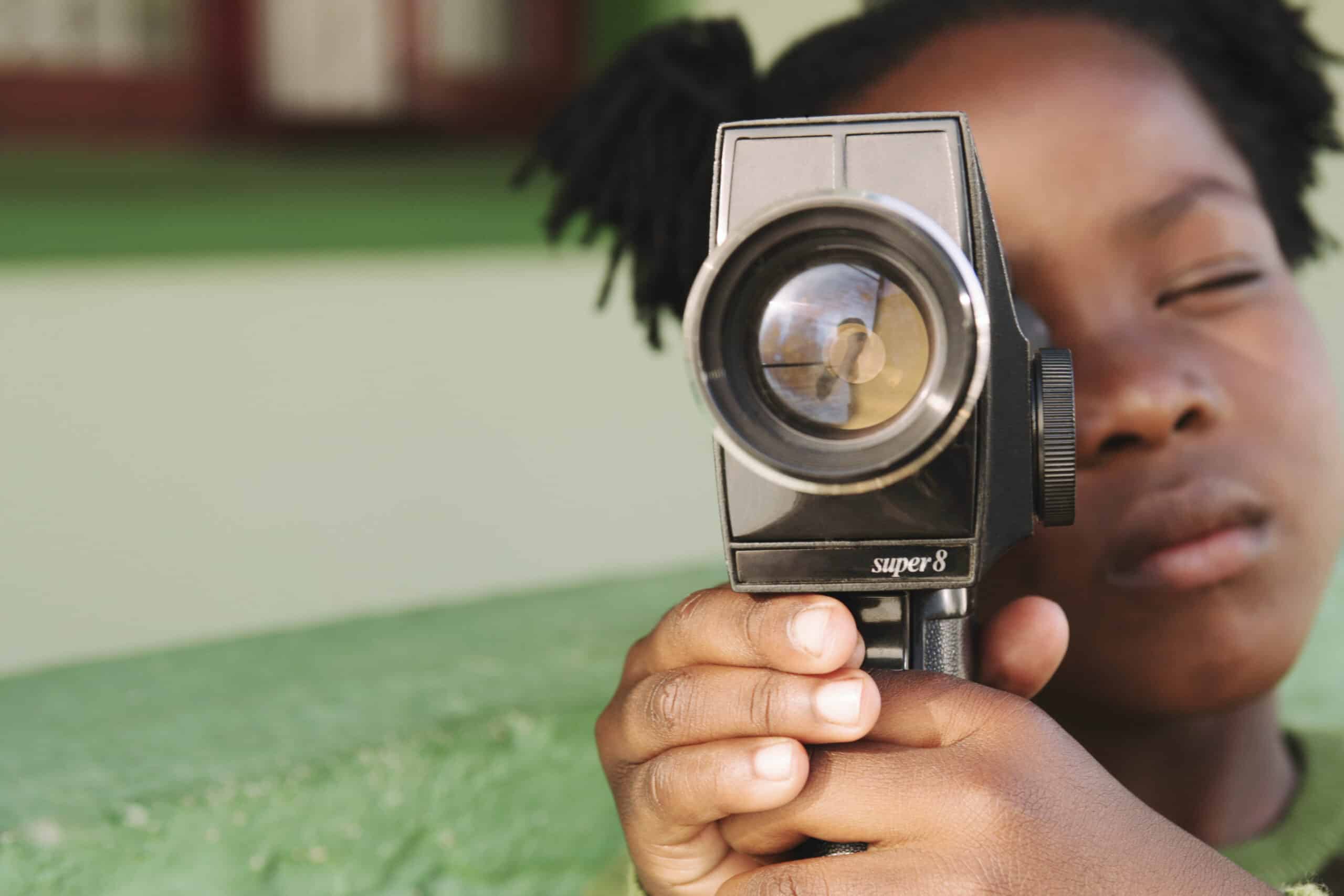 Ein junges Mädchen mit geschlossenen Augen hält eine Super-8-Kamera vor ihr Gesicht. Der Fokus liegt auf dem großen Kameraobjektiv. © Fotografie Tomas Rodriguez