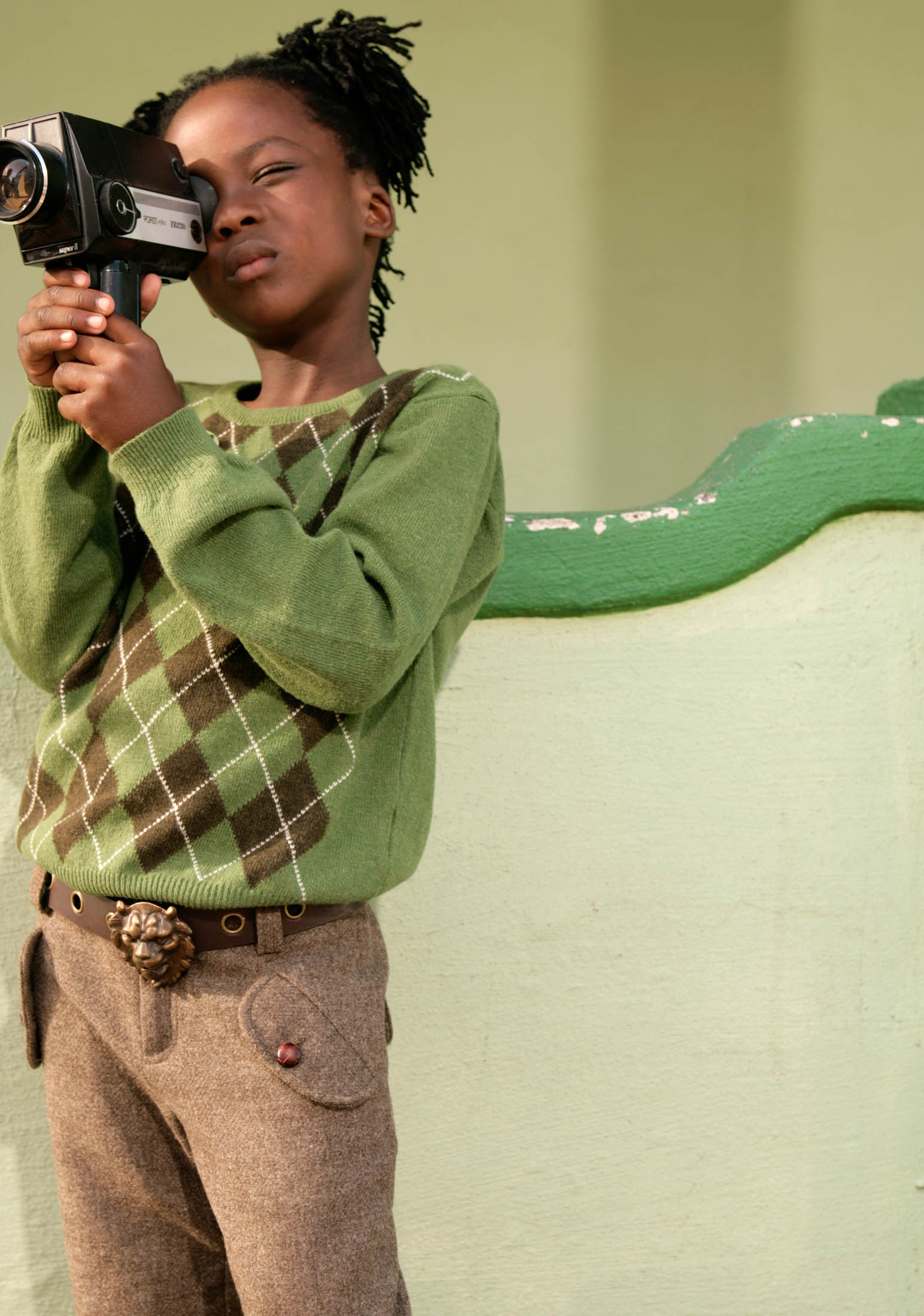 Ein kleines Kind mit geflochtenen Haaren konzentriert sich intensiv darauf, mit einer Handkamera ein Video aufzunehmen. Es trägt einen grün-braunen Pullover und braune Hosen und steht vor einer grünen Wand. © Fotografie Tomas Rodriguez