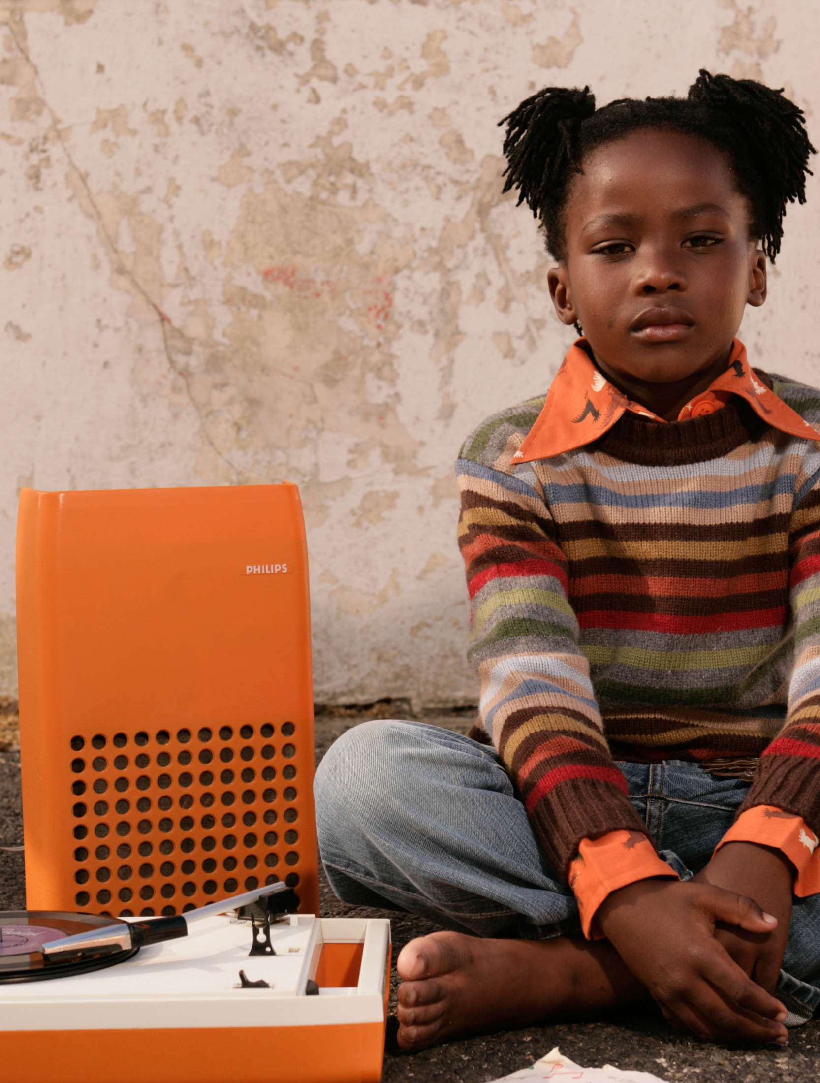 Ein junges Mädchen mit dunkler Haut und hochgesteckten Haaren sitzt auf dem Boden neben einem orangefarbenen Philips-Radio und blickt nachdenklich in die Kamera. Vor ihr liegen eine Schachtel und kleine Gegenstände. © Fotografie Tomas Rodriguez