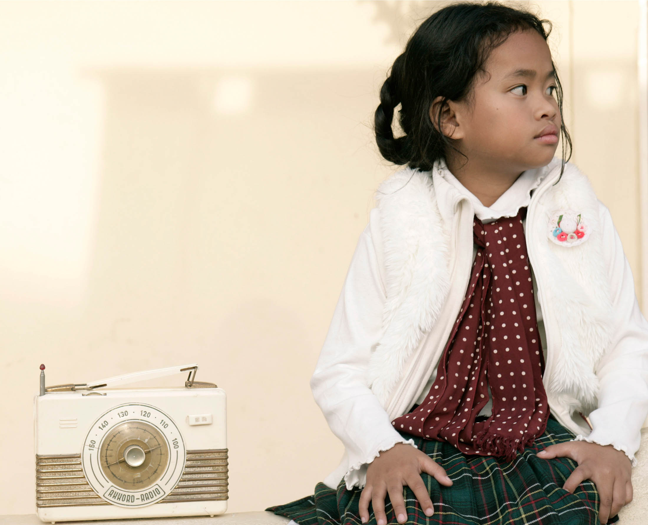 Ein junges Mädchen sitzt neben einem alten Radio und blickt nachdenklich zur Seite. Sie trägt eine weiße Jacke über einer gepunkteten Bluse und einem karierten Rock. © Fotografie Tomas Rodriguez