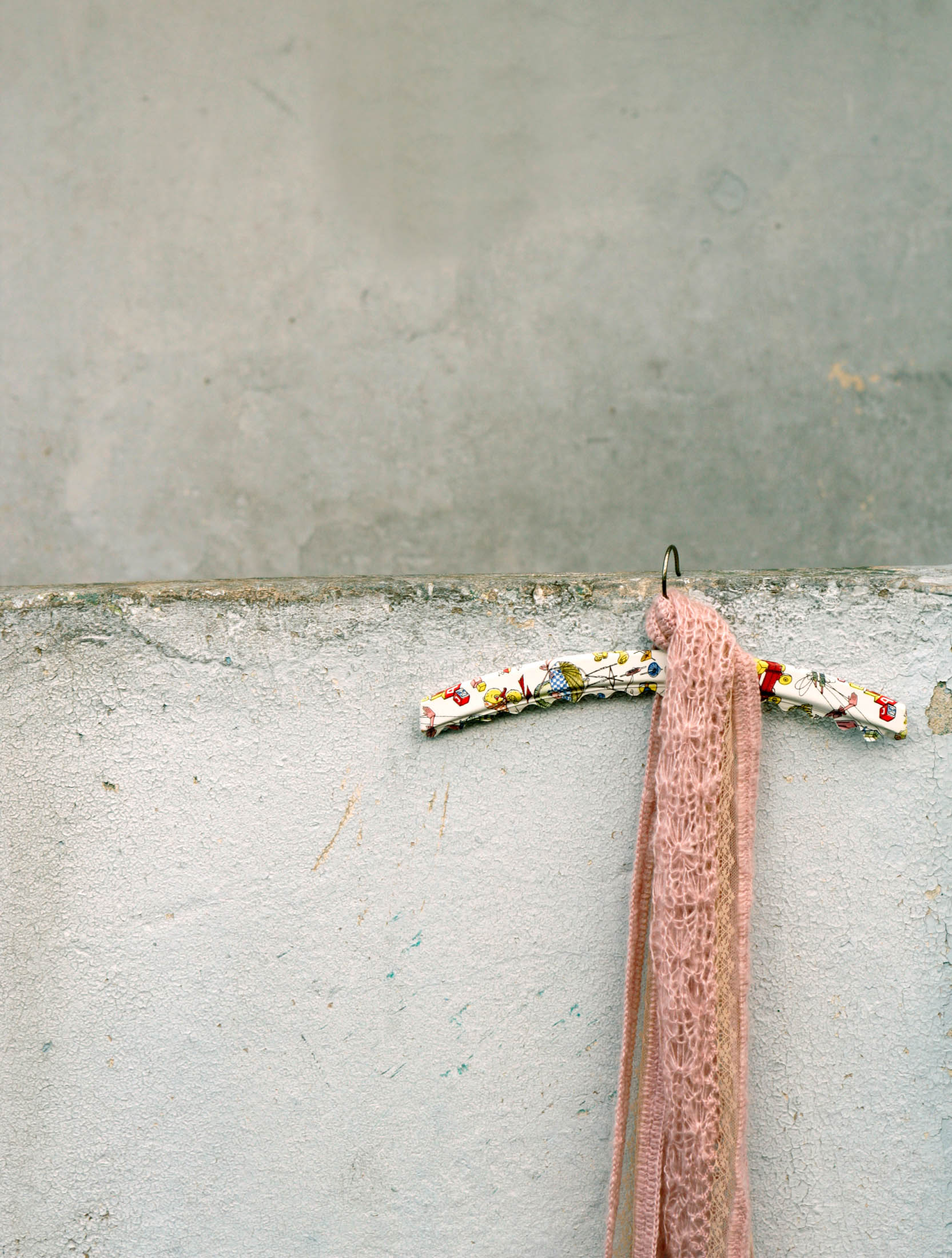 Ein rosa Schal, der auf einem Kleiderbügel mit Blumenmuster vor einer grauen Betonwand hängt. © Fotografie Tomas Rodriguez