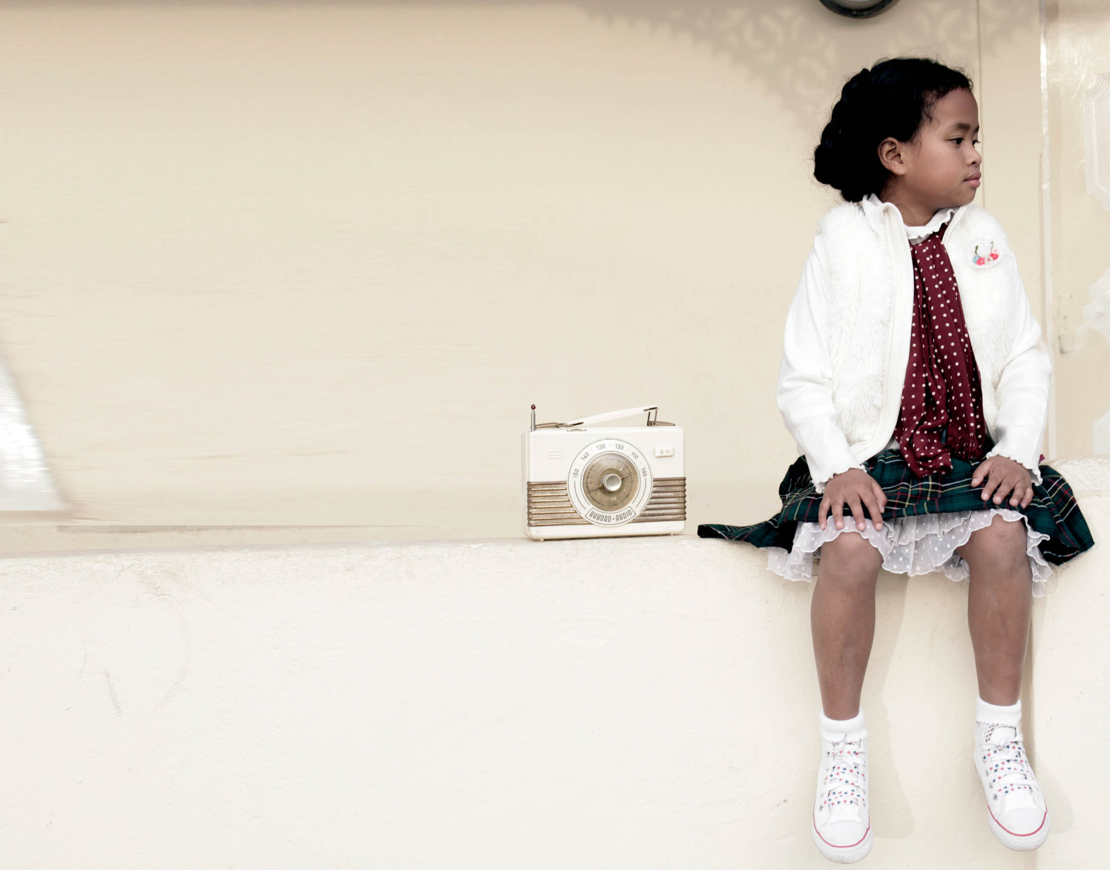 Ein junges Mädchen in weißer Jacke und gemustertem Rock sitzt neben einem altmodischen Radio auf einem Sims und blickt nachdenklich zur Seite. © Fotografie Tomas Rodriguez