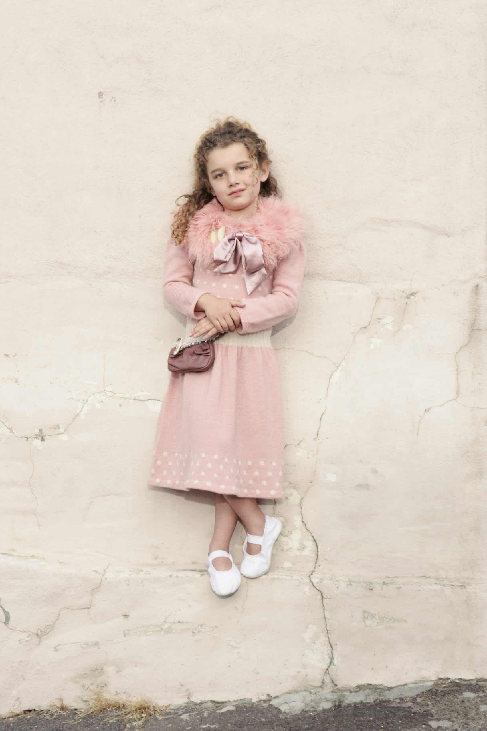 Ein junges Mädchen mit lockigem Haar steht vor einer rissigen Wand. Sie trägt ein rosa Kleid mit Punkten, eine flauschige rosa Jacke und weiße Schuhe. Sie hält eine kleine Handtasche und lächelt leicht. © Fotografie Tomas Rodriguez