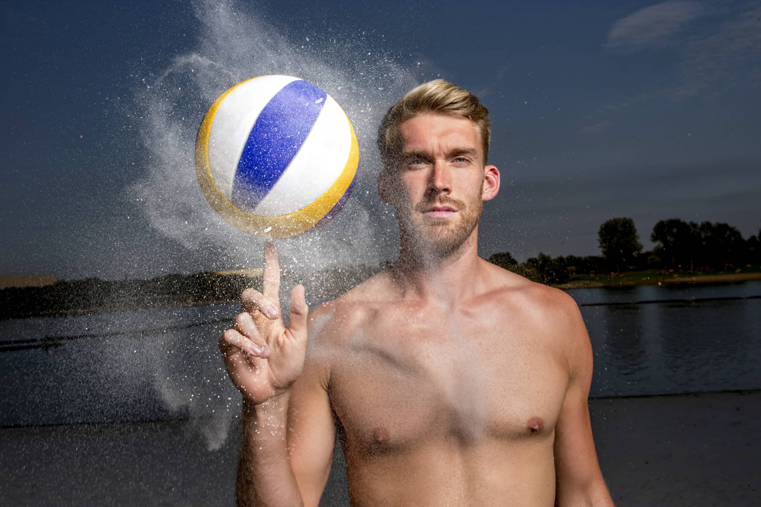 Ein Mann ohne Hemd und mit blondem Haar steht vor einem Gewässer und balanciert einen sich drehenden Volleyball auf seinem Zeigefinger. Der Volleyball hat blaue, gelbe und weiße Abschnitte und etwas Sand fällt von ihm ab. Im Hintergrund sind Bäume unter einem klaren Himmel zu sehen. © Fotografie Tomas Rodriguez
