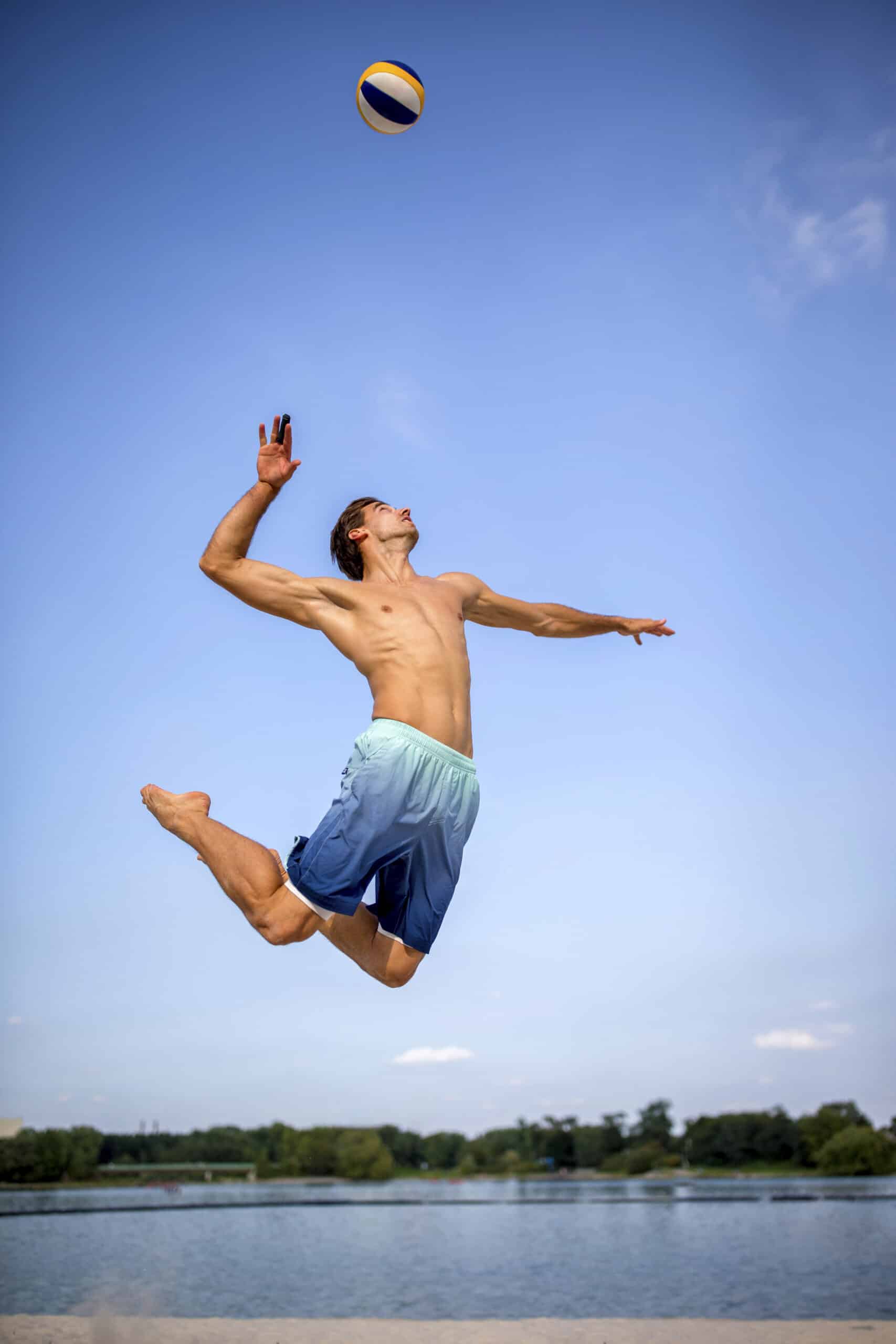 Eine Person in der Luft während eines kraftvollen Volleyballaufschlags an einem Sandstrand. Sie ist oben ohne, trägt blaue Shorts und der Volleyball liegt hoch über ihrem ausgestreckten Arm. Im Hintergrund sind ein ruhiges Gewässer und ein klarer blauer Himmel zu sehen. © Fotografie Tomas Rodriguez