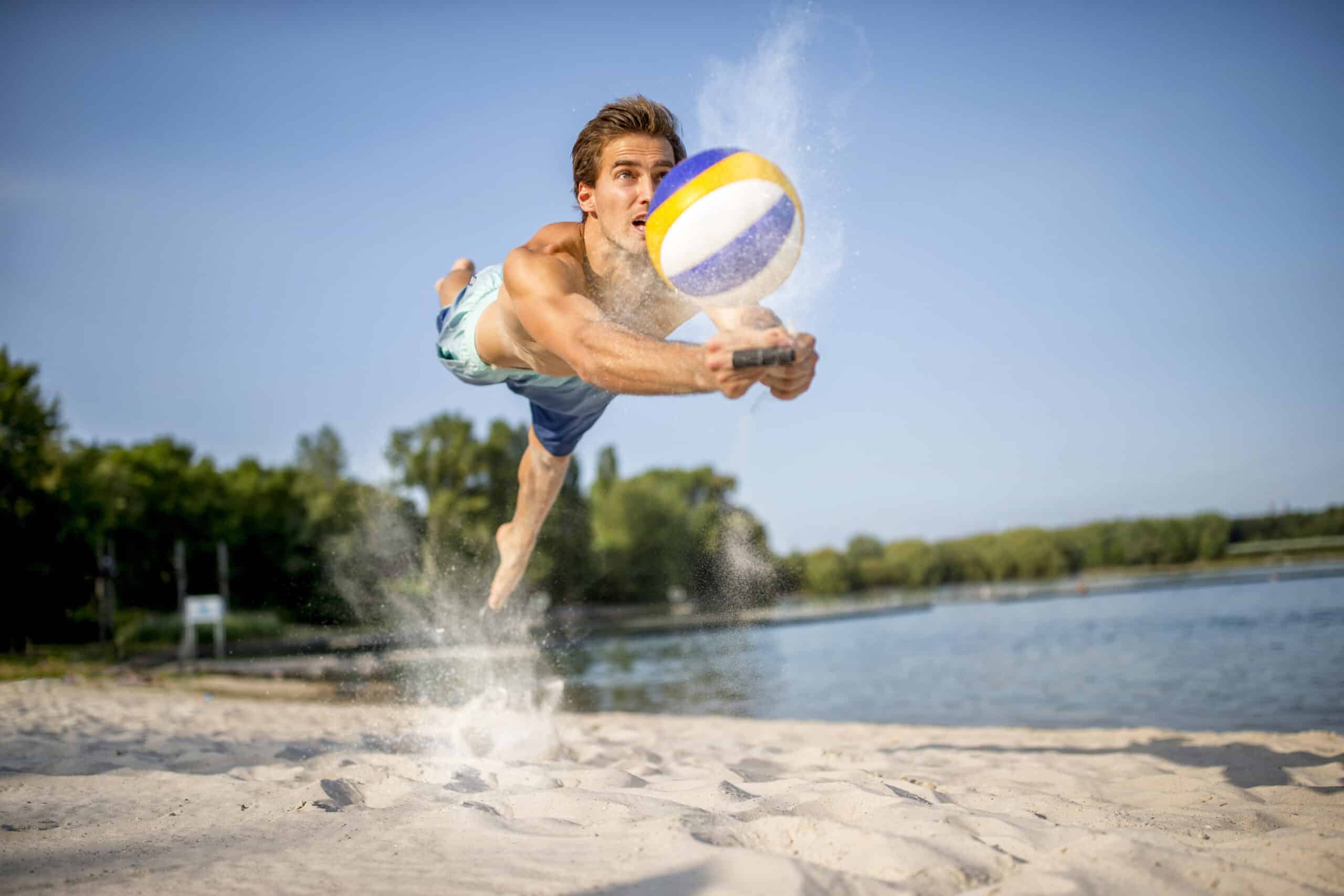 Ein Mann ohne Hemd stürzt sich an einem Sandstrand in die Luft, um einen Volleyball zu schlagen. Der Ball liegt direkt vor seinen Händen und Sand fliegt um ihn herum. Im Hintergrund sind ein Gewässer und Bäume unter einem klaren Himmel zu sehen. © Fotografie Tomas Rodriguez