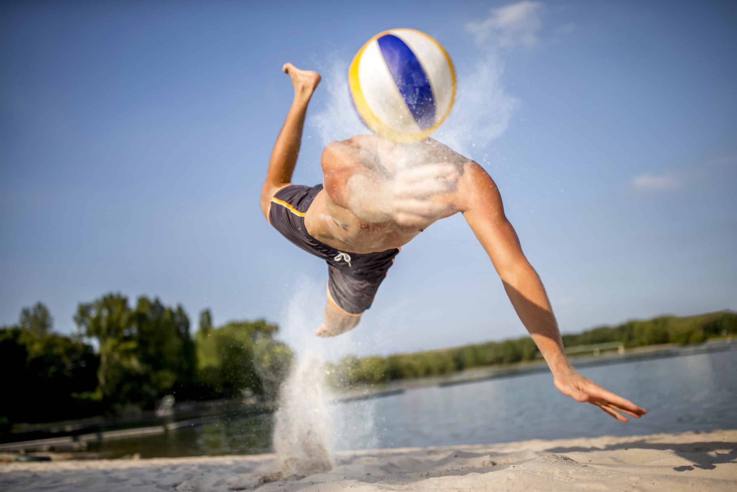 Eine Person ohne Hemd stürzt sich in die Luft, um einen blau-gelben Volleyball an einem Sandstrand zu treffen. Sand wirbelt von der Stelle, an der ihre Füße den Boden verlassen haben. Im Hintergrund sind unter einem klaren blauen Himmel ein Gewässer und grüne Bäume zu sehen. © Fotografie Tomas Rodriguez