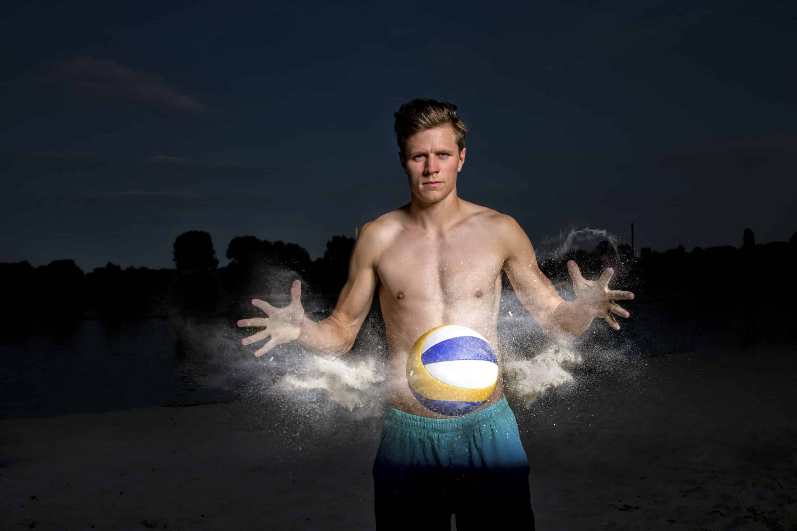 Ein Mann ohne Hemd steht im Freien und scheint einen vor ihm schwebenden Volleyball zu berühren. Seine Hände sind ausgebreitet und eine Wolke aus weißem Pulver umgibt den Ball. Er trägt blaue Shorts und der Hintergrund ist dunkel, was wahrscheinlich auf Abend oder Nacht hindeutet. © Fotografie Tomas Rodriguez
