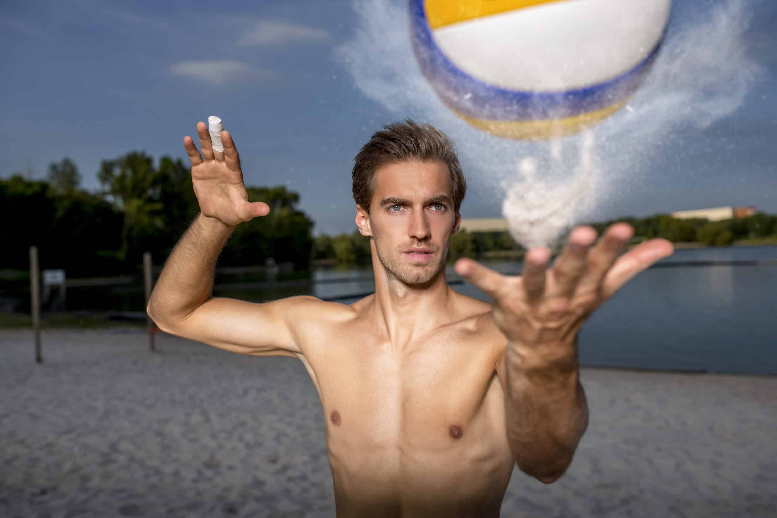 Ein Mann ohne Hemd steht an einem Sandstrand mit Bäumen und einer Wasserfläche im Hintergrund. Er wirft mit einer Hand einen Volleyball in die Luft, während er mit der anderen Hand so kämpft, als würde er ihn gleich treffen. Der Volleyball wird mitten in der Luft gefangen, umgeben von Sandpartikeln. © Fotografie Tomas Rodriguez