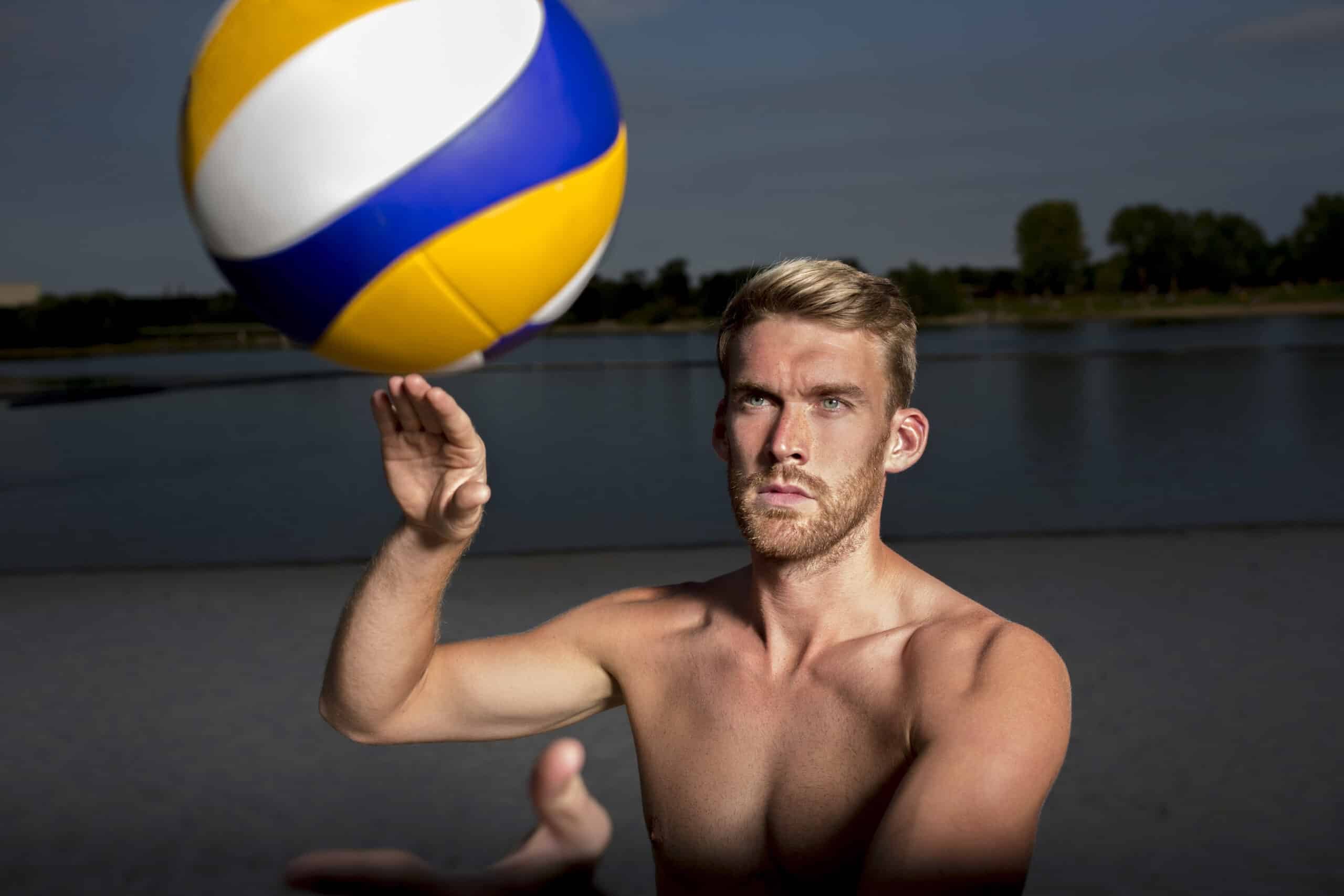 Ein Mann ohne Hemd und mit kurzen blonden Haaren ist gerade dabei, einen bunten Volleyball in die Luft zu schlagen. Er steht im Freien neben einem Gewässer, im Hintergrund sind Bäume zu sehen, der Himmel ist teilweise bewölkt. © Fotografie Tomas Rodriguez